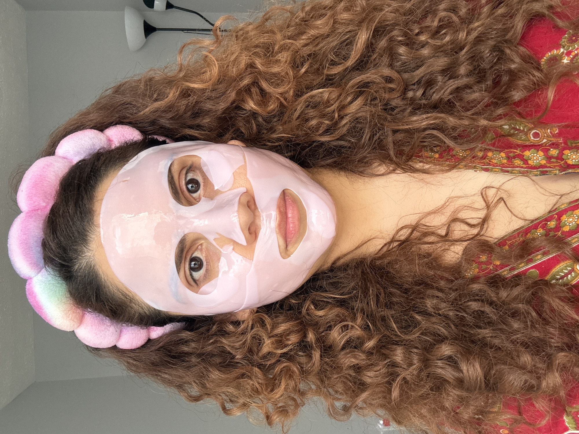 A woman with long, curly brown hair is lying down, wearing a pink sheet mask on her face and a pink and rainbow headband. Her eyes are open, and she is looking at the camera. Part of a patterned red fabric is visible next to her hair. A floor lamp is in the background. The location cannot be determined from the image and is not evident in the image.