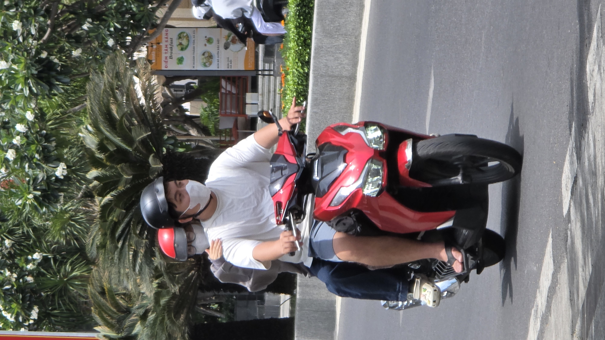 The image depicts two people riding a red and black motorbike. The driver is wearing a white t-shirt, blue shorts, a black helmet, and a white face mask. The passenger, sitting behind the driver, is wearing a red helmet and a white face mask. They are on a paved road, presumably in a city. Adjacent to the road is a sign in Vietnamese and English mentioning "Breakfast" and "Nha Trang Ward". A lush green tree is visible beside the road.