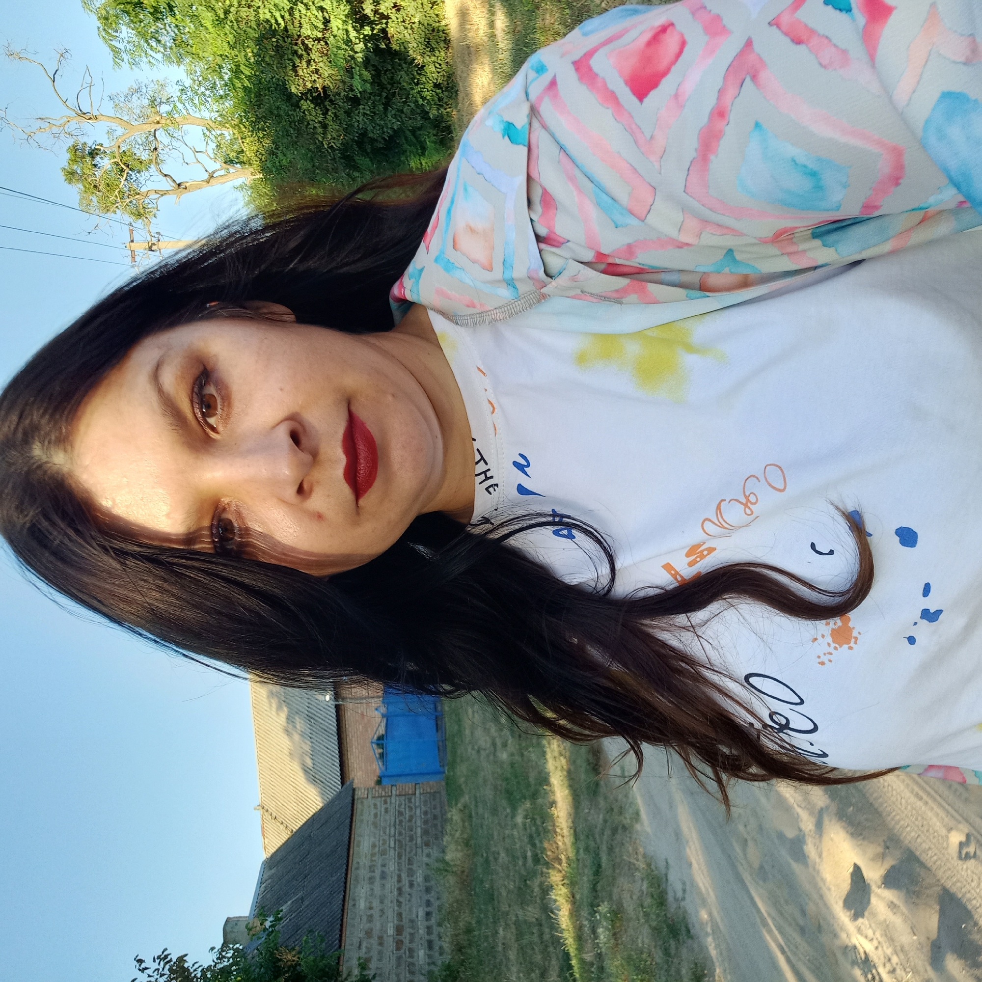 A woman with long, dark hair and dark makeup stands outdoors, facing the camera. She is wearing a white t-shirt with colorful paint splatters and text, and a patterned jacket with pink and blue geometric shapes. The background shows a dirt road, grass, trees, a building with a gray roof, and a blue gate. The lighting suggests it is late afternoon.