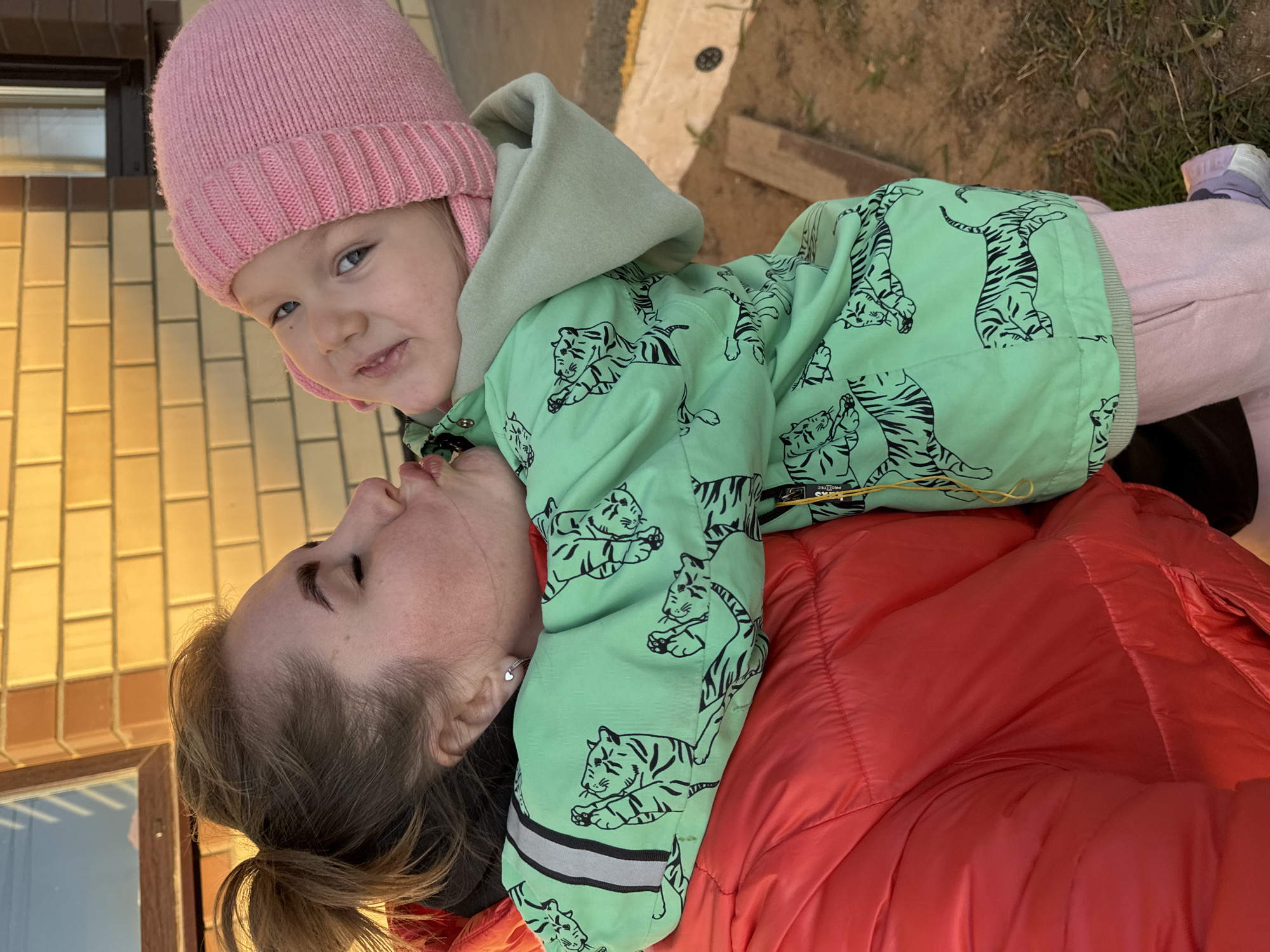 The image shows a young child wearing a pink knit hat and a green jacket with black tiger stripes, lying on top of an adult wearing a red jacket. The child is facing the camera, while the adult's face is turned towards the child. The background includes a building with yellow and brown bricks, as well as some grass and dirt. The location is possibly Ферма, Russia. The interaction appears to be affectionate.