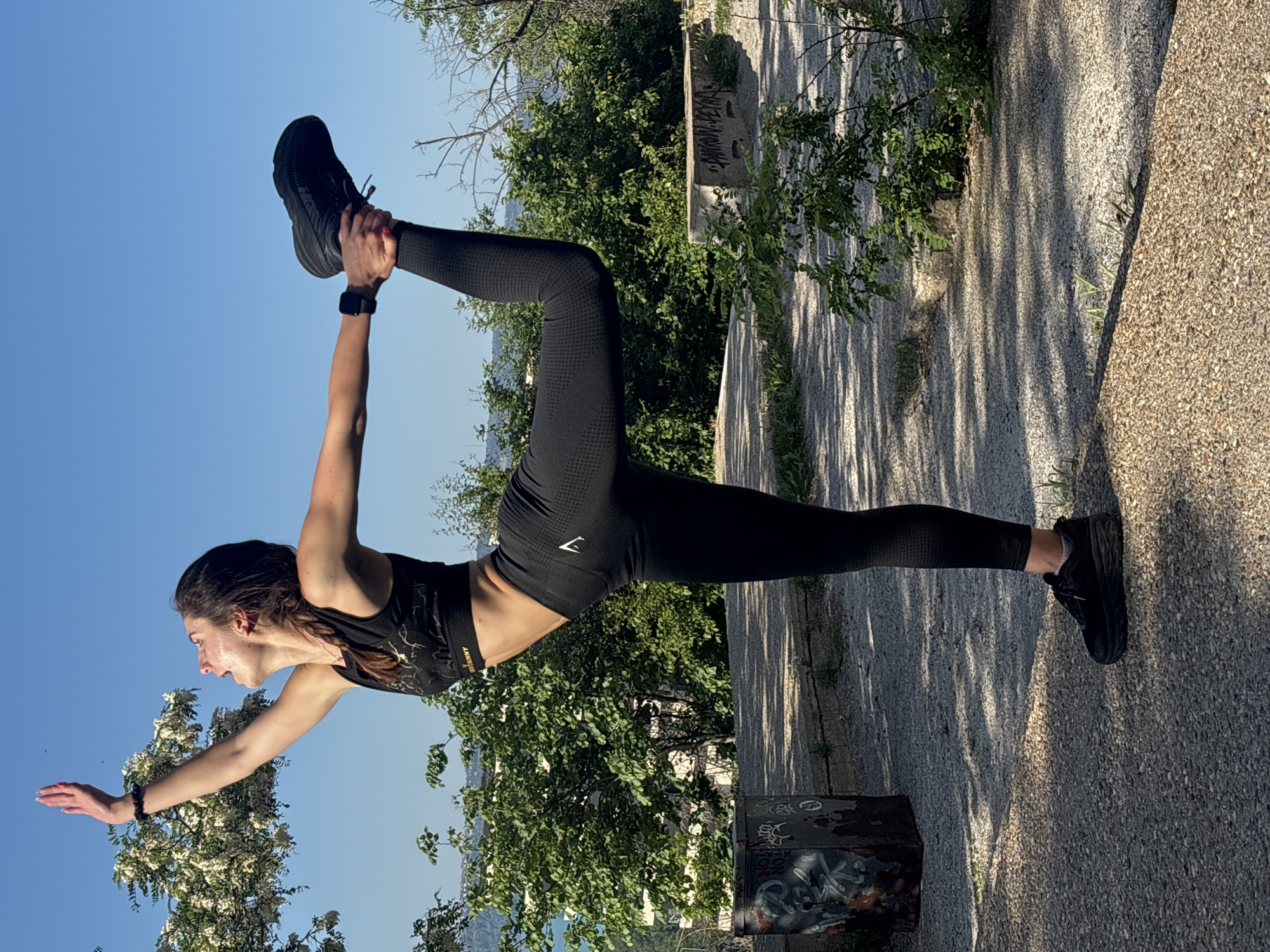 The image shows a woman performing a yoga pose outdoors. She is standing on her right leg, which is grounded on a concrete surface. Her left leg is bent upwards, and she is holding her foot with her left hand. Her right arm is extended forward. She is wearing a black sports bra and leggings. The background consists of trees, a blue sky, and a concrete structure. The image was taken in Varna, Bulgaria. The location seems to be a park or a public space with greenery and concrete elements.