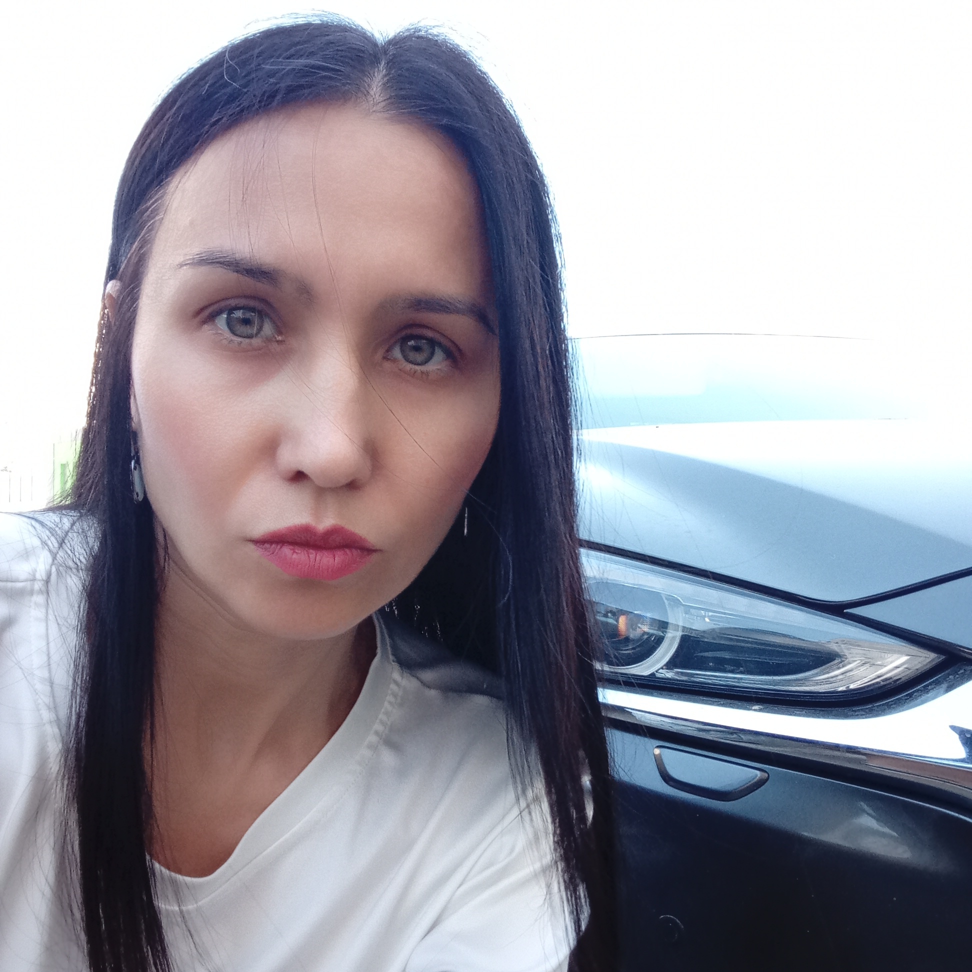 A woman with long, dark hair is positioned in front of a dark-colored car. She is wearing a white t-shirt. The car's front headlight is visible. The setting appears to be outdoors. It is impossible to verify the location as Izhevsk, Russia from the image alone.