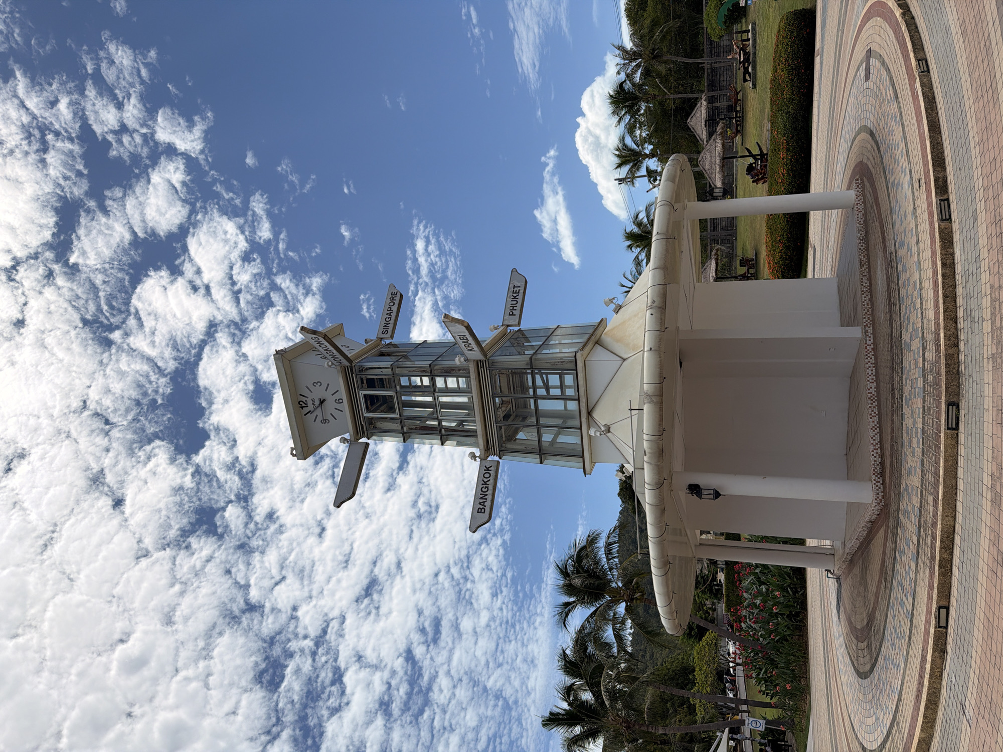 A tall, multi-tiered structure with a clock and directional signs stands in an outdoor plaza on a sunny day. The structure is white and features glass panels on its upper sections. Signs point to various destinations including "BANGKOK," "HONGKONG," "SINGAPORE," "KRABI," and "PHUKET." The plaza is paved with circular patterns of tiles in various colors, and the structure is supported by white columns. Palm trees and lush greenery are visible in the background, along with thatched-roof umbrellas and seating areas in a grassy section. The sky is bright blue with scattered white clouds. The location is the Lamai Beach clock tower in Koh Samui, Thailand. No people are clearly visible or prominently featured in the image.