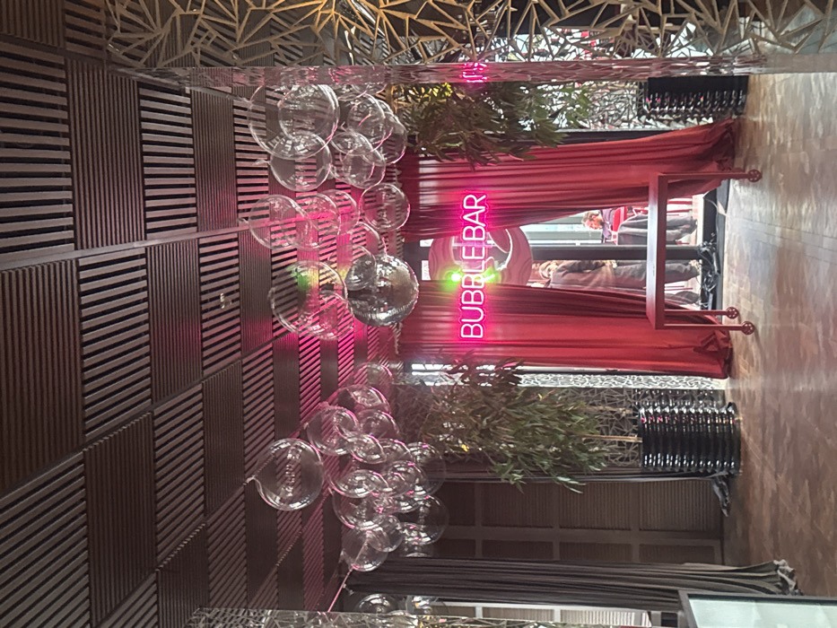 This image depicts the interior of a commercial establishment in Tbilisi, Georgia, specifically an area identified by a prominent neon sign as a "Bubble Bar." The ceiling is constructed from dark wooden slats arranged in parallel patterns, from which numerous transparent glass spheres and reflective orbs are suspended. A vibrant pink neon sign spelling "BUBBLE BAR" is centered above an open archway, which is framed by dramatic, pulled-back red velvet curtains. Two tall, leafy green plants in black, ribbed planters flank the entrance. In the foreground, centered on a light-toned wooden parquet floor, is a rectangular red table equipped with small wheels. Through the archway, two individuals are partially visible: one person stands facing away, wearing a light-colored coat, and another is seated at a table further back, wearing a red top. The right side of the scene features a decorative wall panel with an intricate, geometric, abstract pattern.
