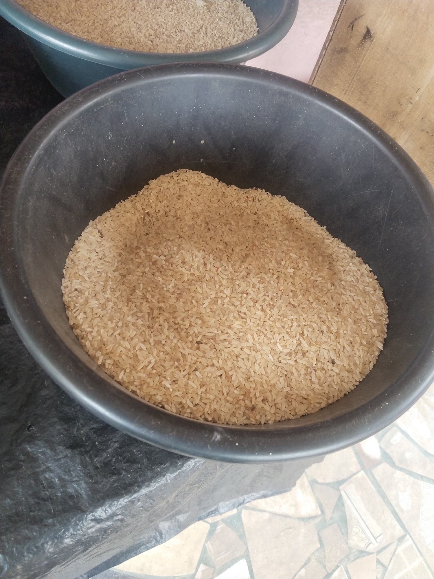 The image shows two large, dark-colored plastic basins filled with uncooked rice. The basin in the foreground is larger and more prominently displayed, with its rim clearly visible and a substantial amount of rice filling it. The rice appears to be light brown or yellowish in color and is piled high, with individual grains distinguishable. The basin is sitting on a black, crinkled surface, possibly a tarp or plastic sheet. Behind this basin, and slightly out of focus, is another, similar basin, also containing rice. This second basin appears to be a darker teal color. The background is blurred, but hints of a wooden surface and a tiled floor can be discerned. The overall impression is one of bulk storage of rice, likely in a market or storage facility. The image's context suggests it was taken in Uyo, Nigeria, an environment where such storage of staple food items is common. There are no people visible in the image.