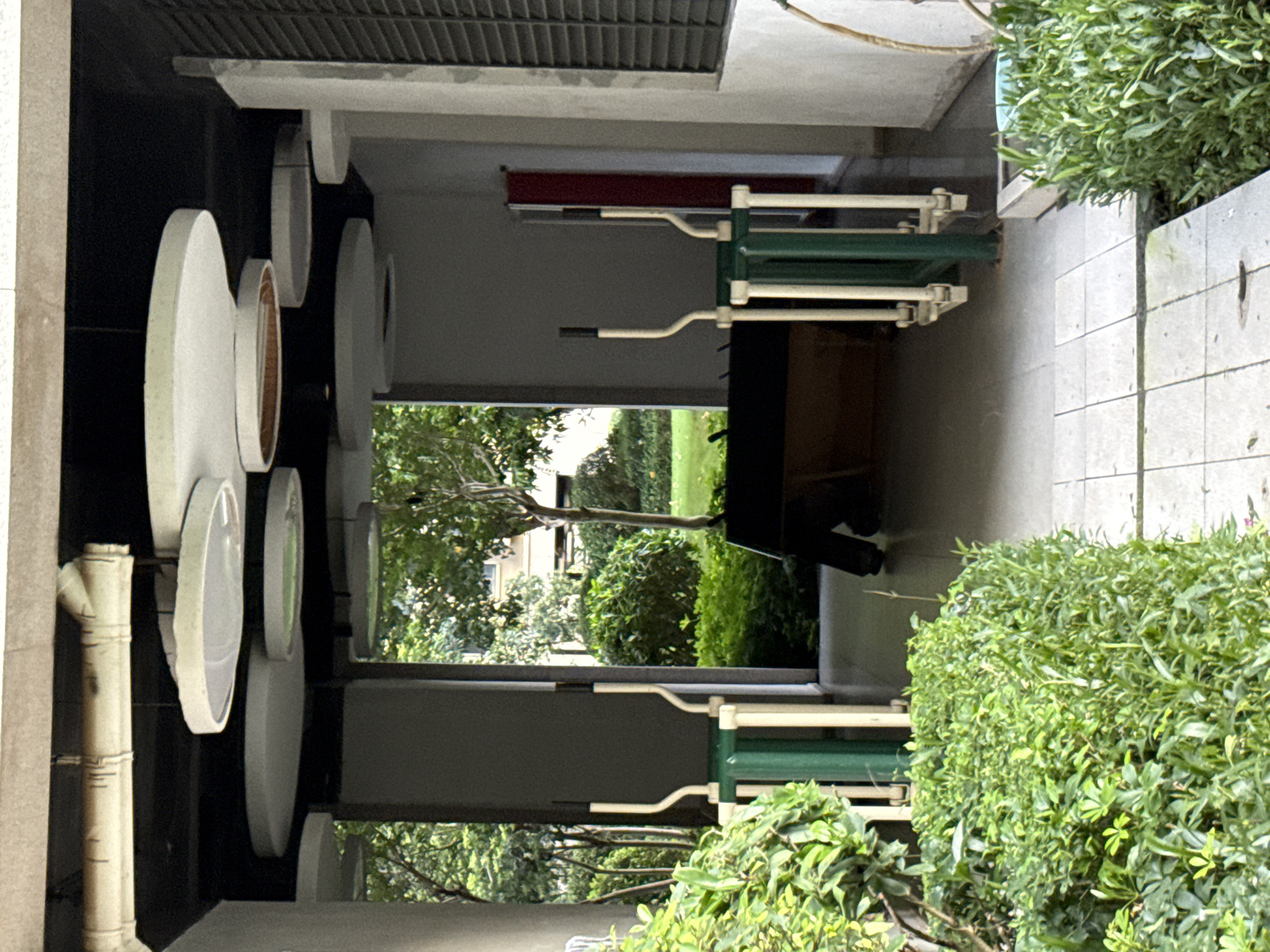 The image depicts an outdoor covered walkway area with exercise equipment and lush greenery. Two elliptical trainers, one on each side of the walkway, are positioned centrally. The trainers are primarily white with green vertical supports. In the background, through an open archway, a verdant garden with trees and bushes is visible, along with the facade of a building. Overhead, circular decorative light fixtures are suspended from the ceiling. The walkway itself is paved with light-colored square tiles. The scene appears to be a communal space within a residential or recreational area. No individuals are clearly visible, although legs of a person are partially obscured in the background, near a dark rectangular object. The setting is likely in Huizhou, China, given the context.