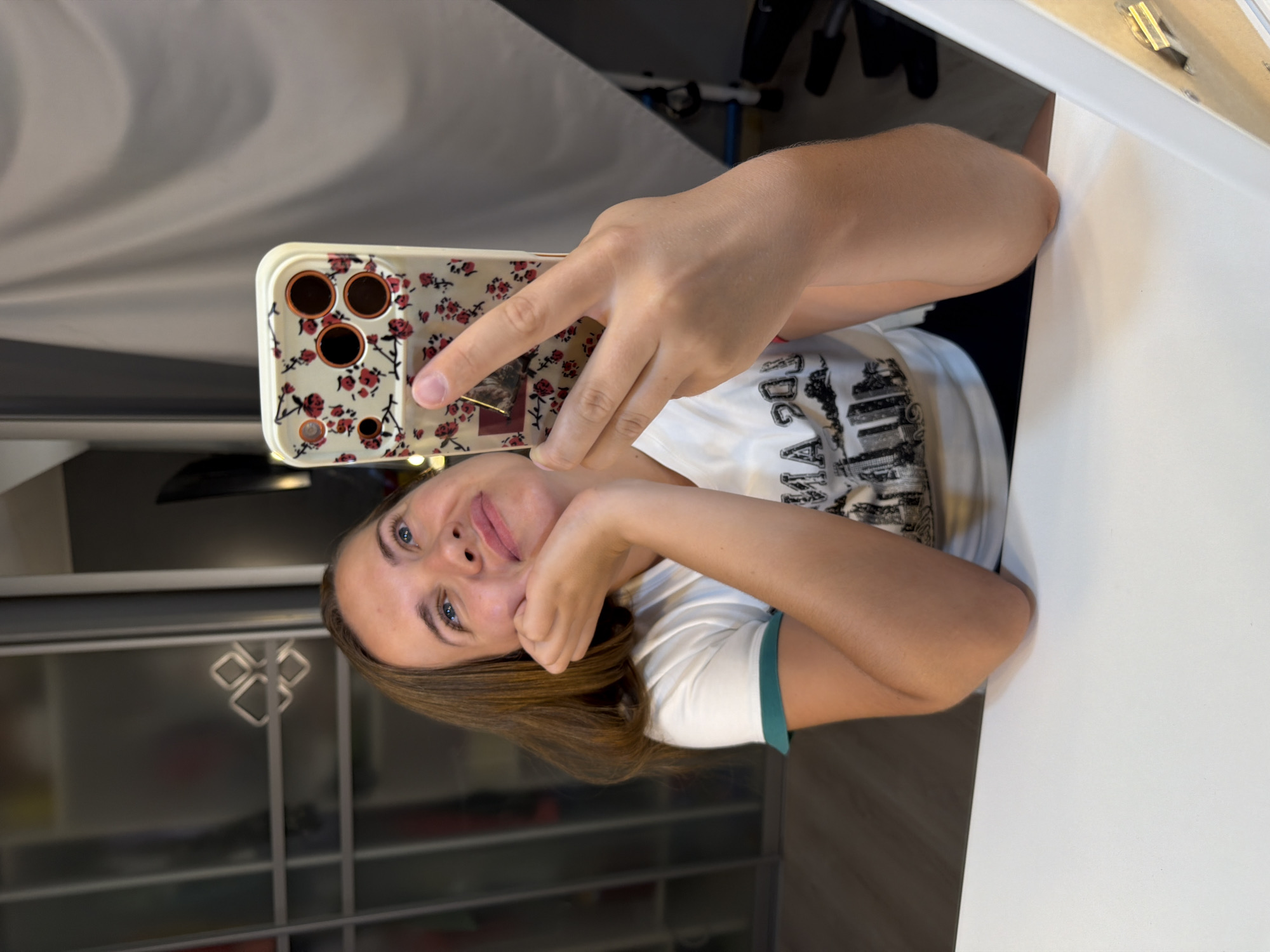 A young woman with shoulder-length brown hair, wearing a white t-shirt with a graphic print and a teal collar, is taking a selfie with her smartphone. She is leaning on her hand, looking upwards with a thoughtful expression. The phone has a floral pattern case with red flowers. The background shows a room with frosted glass cabinets and a white curtain. The scene does not provide any information about the city of Sochi, Russia.