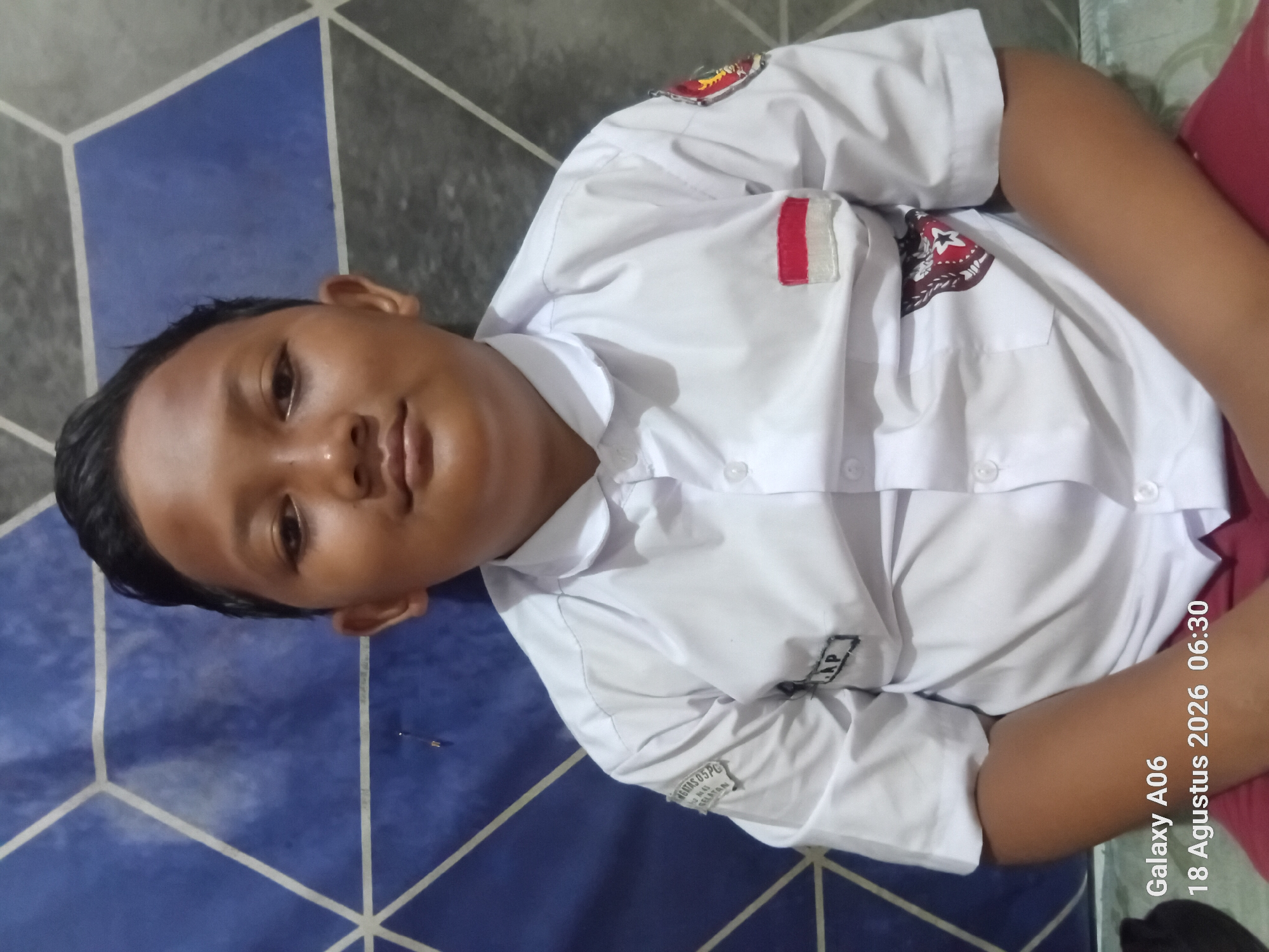 This is a media file of a young boy lying down on a blue tiled floor, wearing a white school uniform with a red and white flag patch on his left chest and a school emblem on his right chest. The uniform has a collar and buttons down the front. There is some text visible on the uniform, including "AP" on a small patch. The boy's face is turned towards the camera, and he appears to be relaxed. A red garment is visible behind him. The image also contains text overlay from a phone, indicating it was taken with a "Galaxy A06" on "18 Agustus 2026 06:30". The scene appears to be indoors, and the time suggests it is early morning. The location context is South Jakarta, Indonesia.