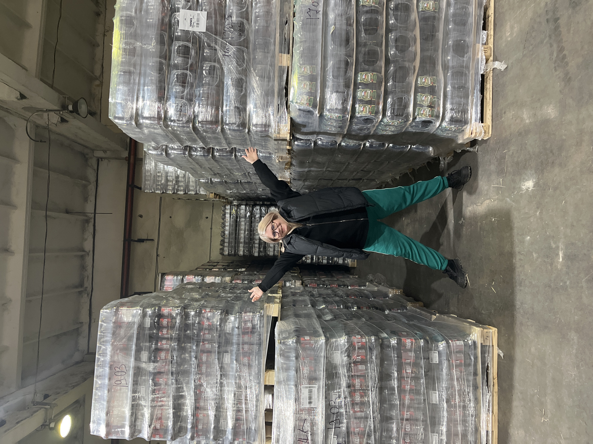 The scene depicts the interior of a large industrial warehouse or storage facility in Izhevsk, Russia. A middle-aged woman with blonde hair and glasses stands centrally between two towering stacks of palletized goods. She is wearing a black puffer vest over a black long-sleeved shirt, green sweatpants with a white stripe, and black sneakers. Her arms are outstretched horizontally to her sides, with hands open, and she is smiling directly at the camera.
The warehouse features a concrete floor with minor debris and scuff marks, and a concrete ceiling with exposed structural beams, utility pipes (including a red pipe segment), and industrial lighting fixtures, some of which are illuminated.
The goods are predominantly beverages, specifically alcoholic products, tightly shrink-wrapped in clear plastic and stacked on wooden pallets. The stack on the left displays numerous packages of "OXOTA" (ОХОТА) branded products, identifiable by red and dark brown cans. The stack on the right includes some packages with green branding, likely another type of beer. Handwritten labels and alphanumeric codes, such as "19.03", "12425673", and "170", are visible on the plastic wrapping of various pallets. Additional stacks of goods are visible in the darker background beyond the central subject.