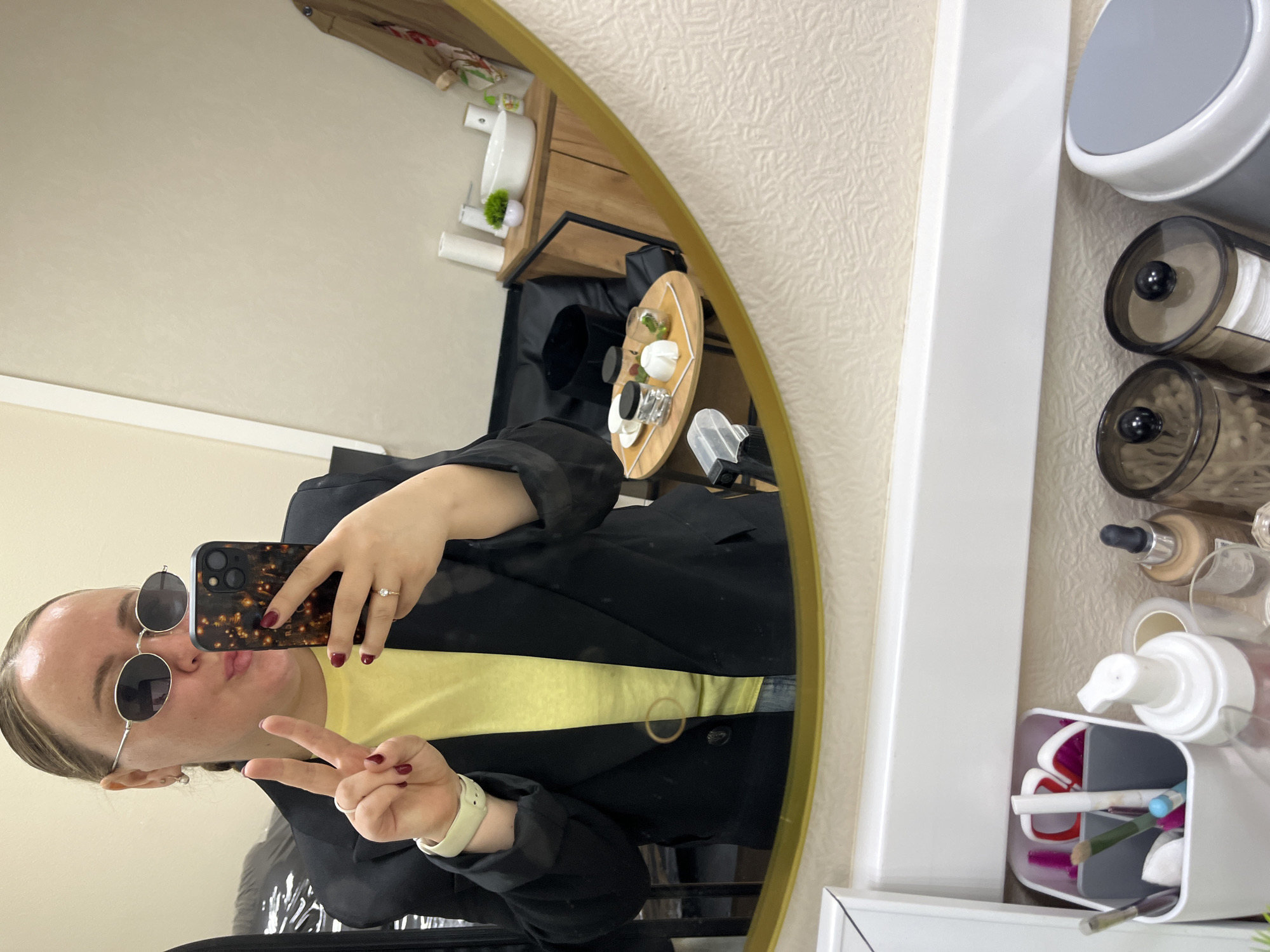 The image shows a person taking a selfie in a mirror. The person is wearing sunglasses, a yellow shirt, and a black blazer. They are holding a phone in their right hand and making a peace sign with their left hand. The background includes a shelf with various items, including a container with cotton swabs, bottles, and a makeup organizer. The mirror has a gold frame. The location is potentially Chaikovsky, Russia, based on prior knowledge.