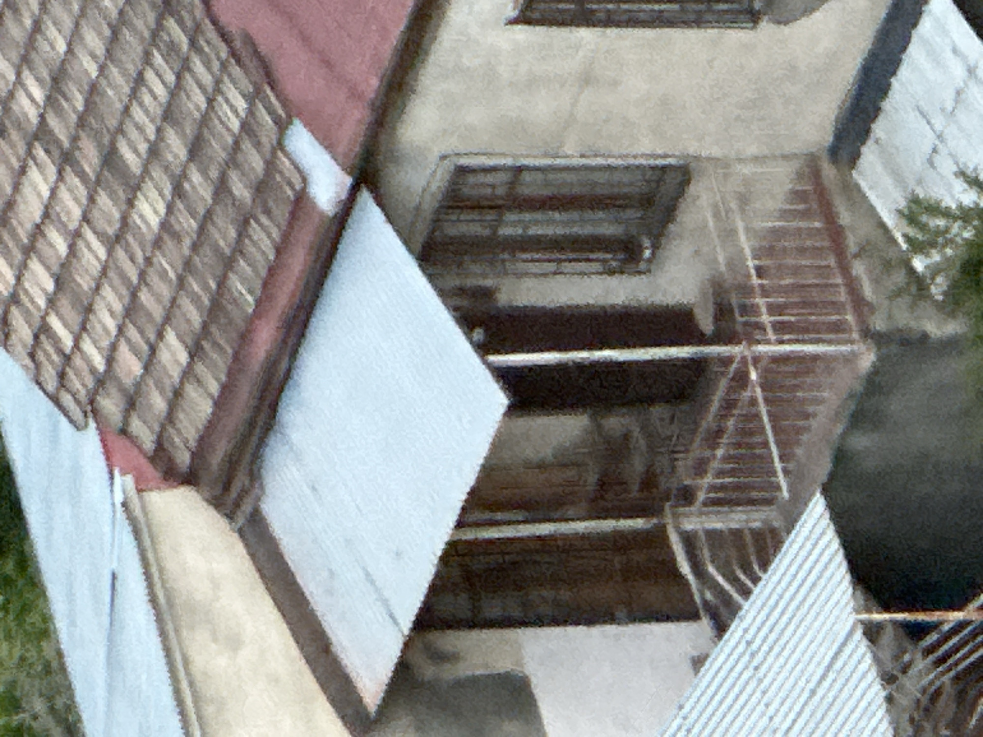 This high-angle, possibly aerial, shot captures a detailed view of several interconnected rooftops and building sections, likely within an urban environment such as Tbilisi, Georgia. The perspective is tilted, emphasizing the geometric patterns and textures of the architectural elements.
In the upper left, a prominent roof features reddish-brown, possibly terracotta, tiles arranged in a diagonal grid pattern. Adjacent to this, and extending towards the center, are sections of light-colored concrete or stucco surfaces, suggesting flat rooftops or terraces. A section of light blue or grey corrugated material, possibly a metal roof, is visible in the mid-left and again in the lower right, showing distinct diagonal ridges.
Within the scene, several structural details are apparent. In the upper middle, a dark, horizontal structure resembling an awning or balcony can be seen above what might be a window. To its right, a set of metal stairs with railings descends, hinting at an exterior access point between building levels. Other metal frameworks, possibly railings or decorative elements, are scattered throughout the image, particularly in the lower right and central areas.
No people are visible, and no specific activity is taking place. The clear and bright lighting suggests it is daytime with good weather. Small patches of green foliage are visible at the edges of the image, indicating some surrounding vegetation. There is no visible text.