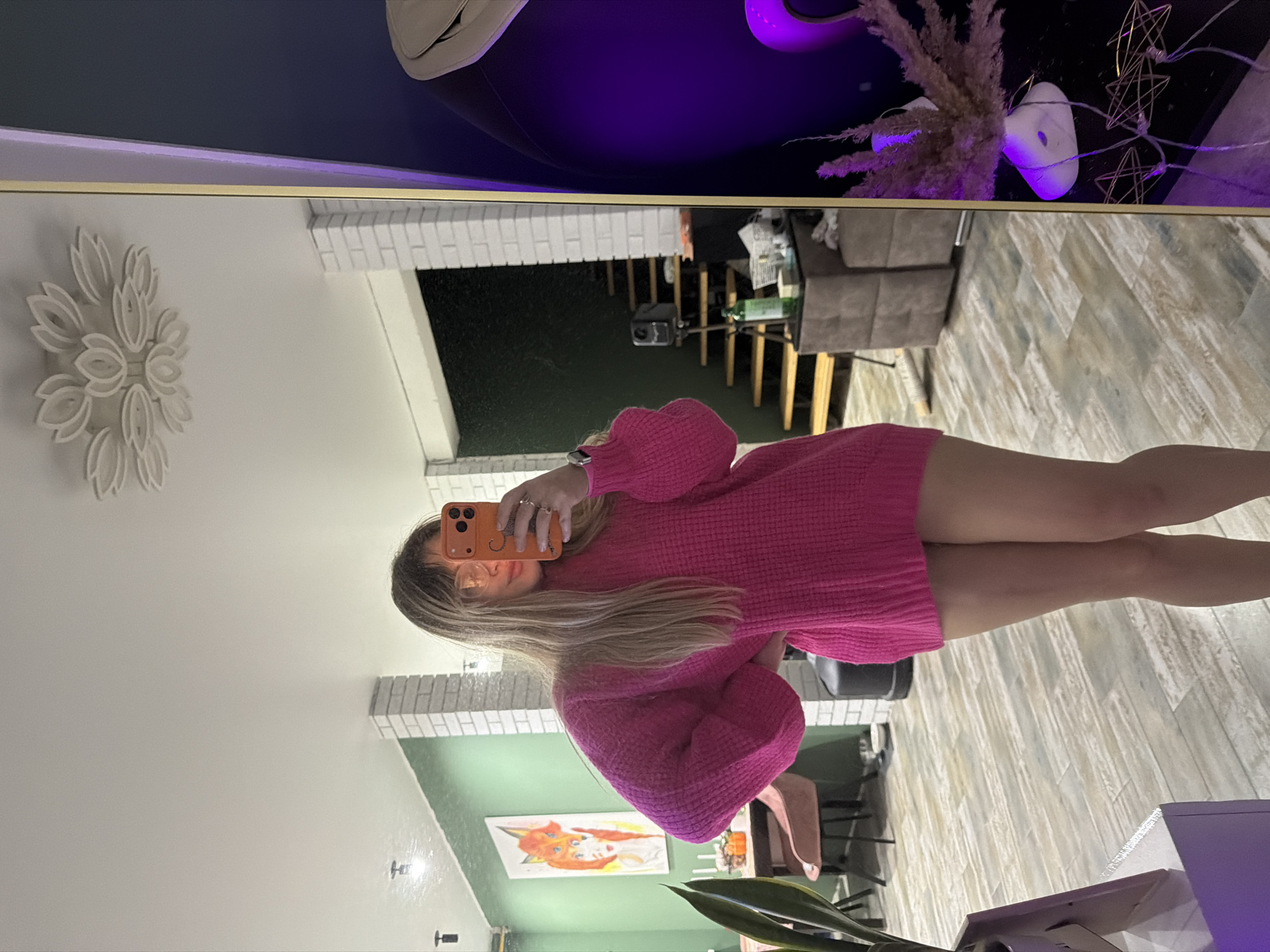 A person takes a selfie in a mirror. The person is wearing a bright pink, sweater dress. The mirror reflects the surrounding room, which includes a staircase, artwork, furniture, and decorative items. The location may be in the city of Большая Шильна, Russia.