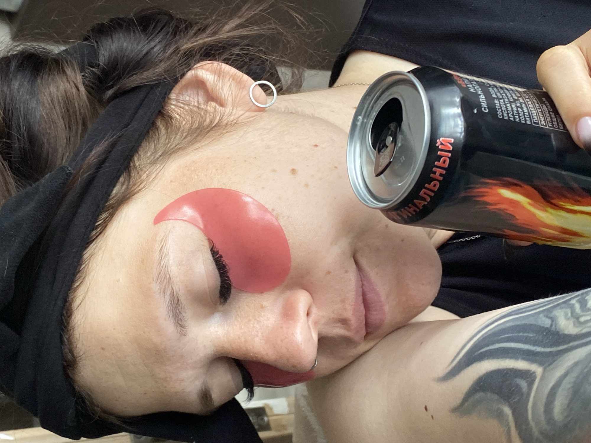 A woman with closed eyes and pink under-eye patches rests her head on her shoulder, holding a can of energy drink. She has a black headband on and her hair is tied up. She is wearing a black shirt and a silver hoop earring. The energy drink can has flames on it and text in Russian. A tattoo is visible on her arm. The background is blurry, but appears to be indoors with some light filtering in. The city of Chaikovsky, Russia, is not identifiable in the image.