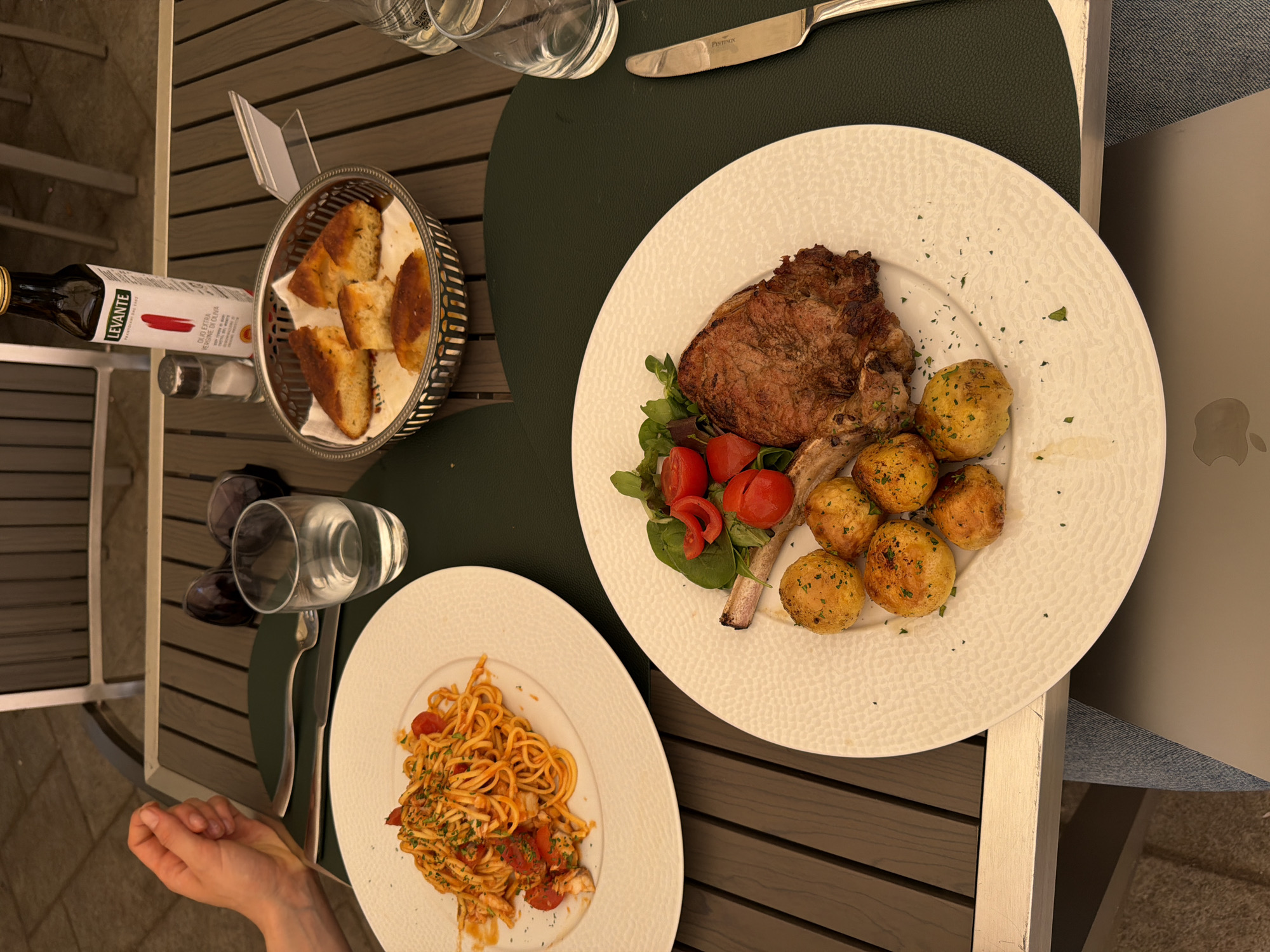 A high-angle, medium shot captures a table setting for two at an outdoor dining establishment in Milan, Italy. The table, made of dark wood planks, is covered in part by a dark green placemat. On the table, two white, textured plates are visible. The plate on the left contains a serving of spaghetti with a red sauce, garnished with herbs and cherry tomatoes. The plate on the right features a grilled pork chop, accompanied by roasted potatoes and a small salad of mixed greens and cherry tomatoes. In the background, a silver basket holds several pieces of toasted bread, and a bottle of Levante Extra Virgin Olive Oil, a salt shaker, and a glass of water are also present. A pair of sunglasses rests on the table near the glass. The scene suggests a leisurely meal in a city known for its culinary scene, possibly in a restaurant with an outdoor seating area in Milan, a fashion and design capital of the world.