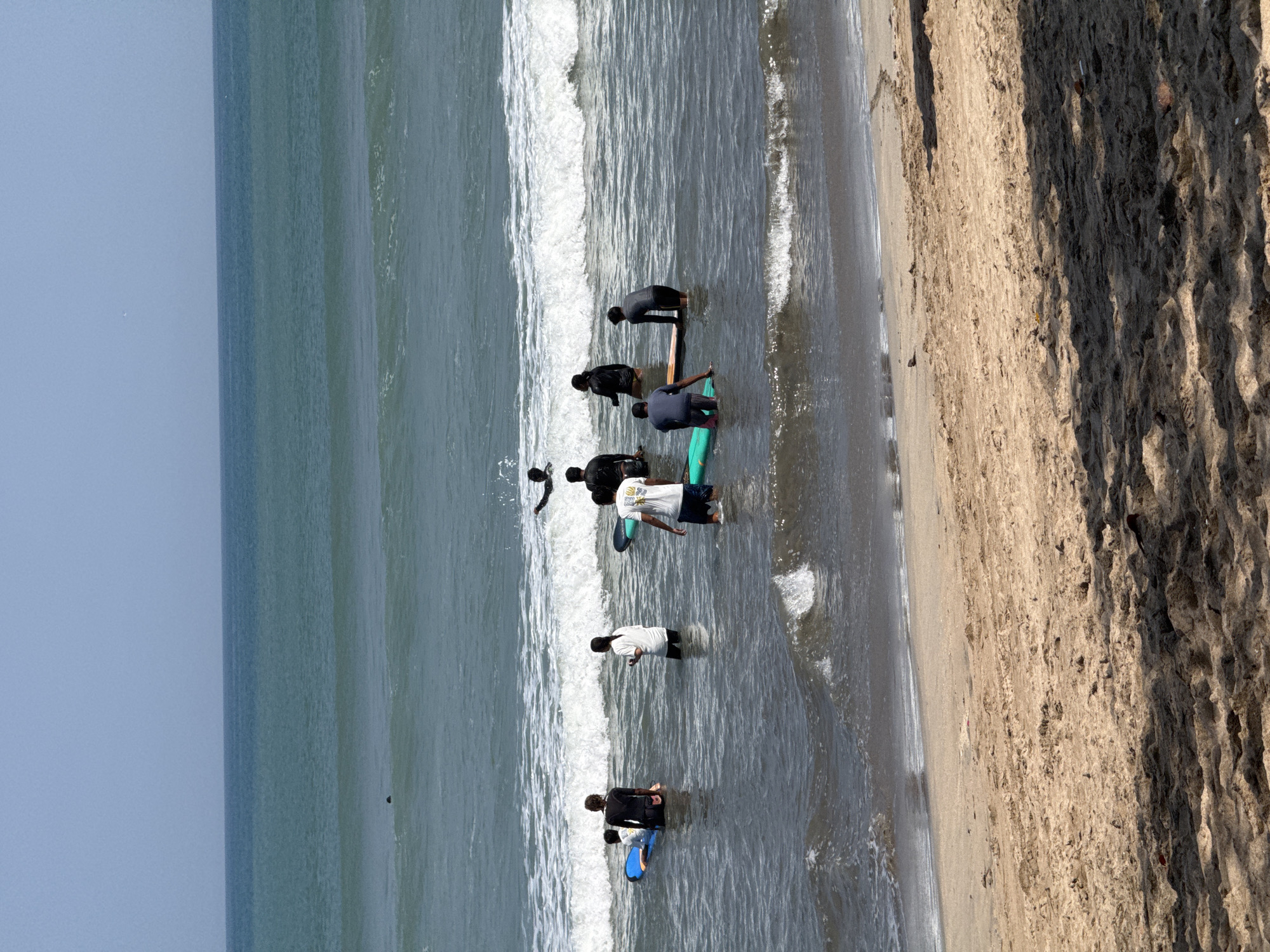 In Kuta, Indonesia, at a sandy beach with gentle waves, a group of approximately eight individuals are engaged in a surfing lesson. Seven people are in the water, some with surfboards. One individual, wearing a white t-shirt and blue shorts, stands in the water, facing away from the camera. Another person, in dark clothing, is crouched on a surfboard. Two individuals in dark wetsuits are further out in the water, one standing and the other partially submerged with a surfboard. A person in a white t-shirt and black leggings stands in the water, while another in a black wetsuit is positioned on a surfboard to their left. To the far left, a person in a dark wetsuit is on a blue surfboard. In the background, a person in black is on a surfboard further out in the ocean. The sky is clear and pale blue. The sand in the foreground is light brown, with shadows cast by unseen objects.