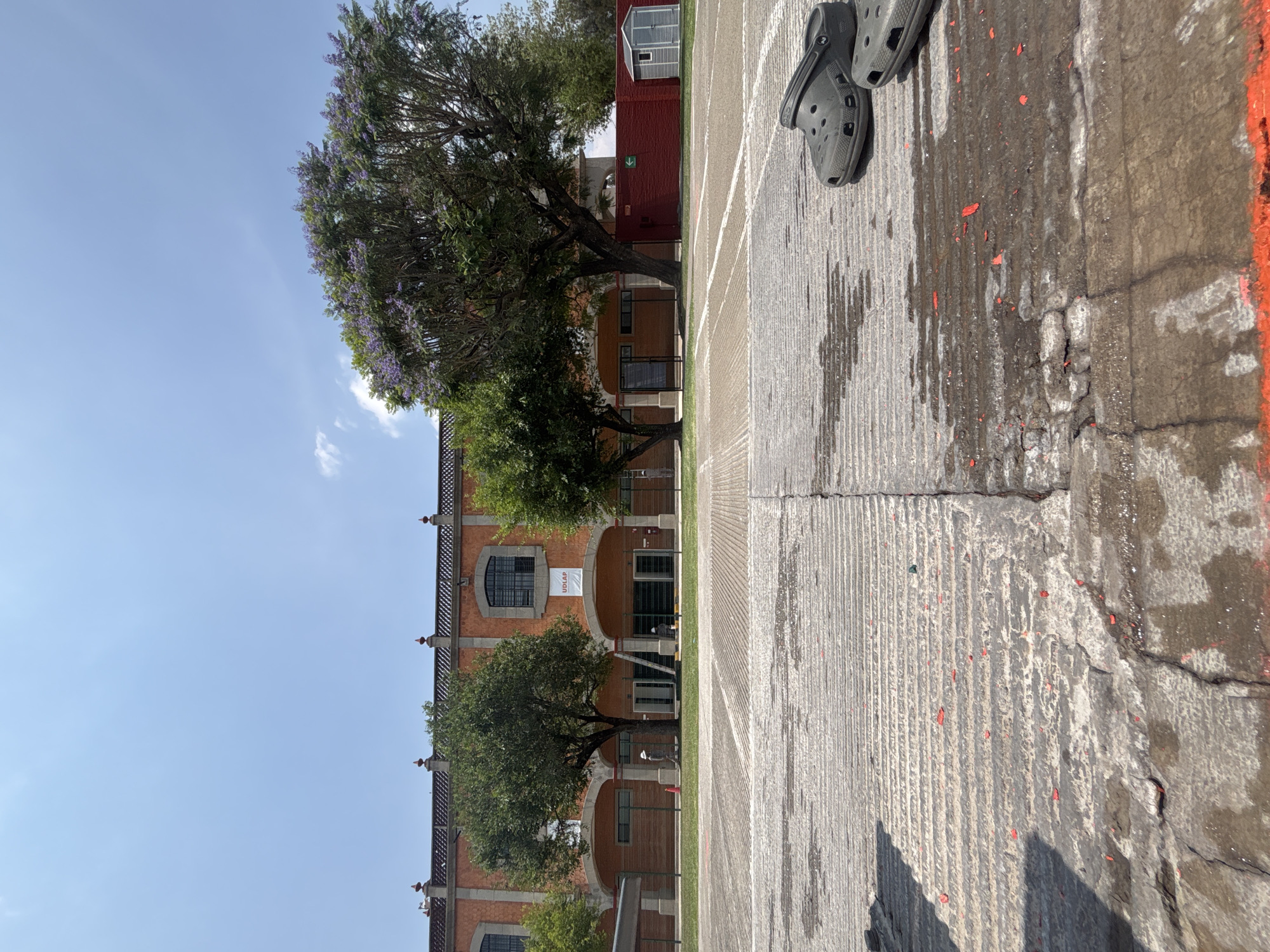 The image captures a scene in San Andrés Cholula, Mexico. A brick building with arched windows is partially visible, featuring a sign that reads "UDLAP". Several trees are growing in front of the building. The foreground consists of a cracked concrete surface with white and orange markings. A pair of gray Crocs sandals sits on the edge of the concrete. The sky is blue with few clouds. There are two people standing by the building under construction.