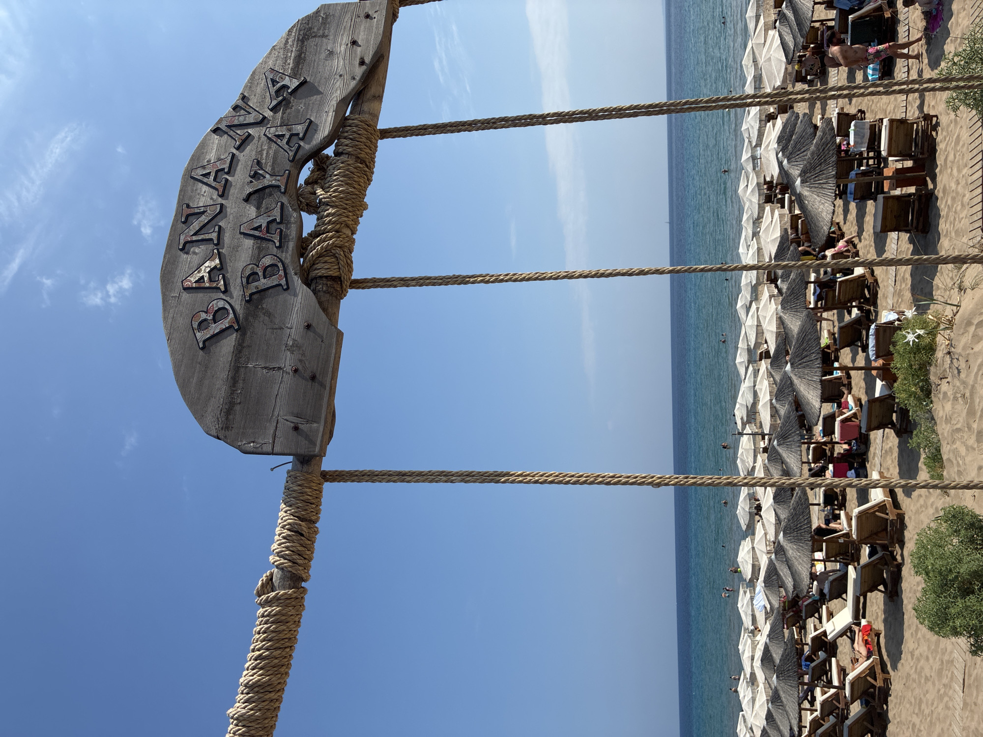 The image depicts a beachfront scene at Banana Beach, Vasilikos, Zakynthos, Greece, under a clear blue sky. In the foreground, a weathered wooden sign with the words "BANANA BAY" in textured letters hangs from thick ropes. Below the sign, rows of white and dark straw umbrellas cover numerous beach chairs and loungers arranged on the sandy beach. People are visible relaxing on the loungers, some with umbrellas providing shade. The turquoise ocean stretches to the horizon, with a few people swimming or wading in the water. The overall atmosphere is one of a busy, popular beach resort.