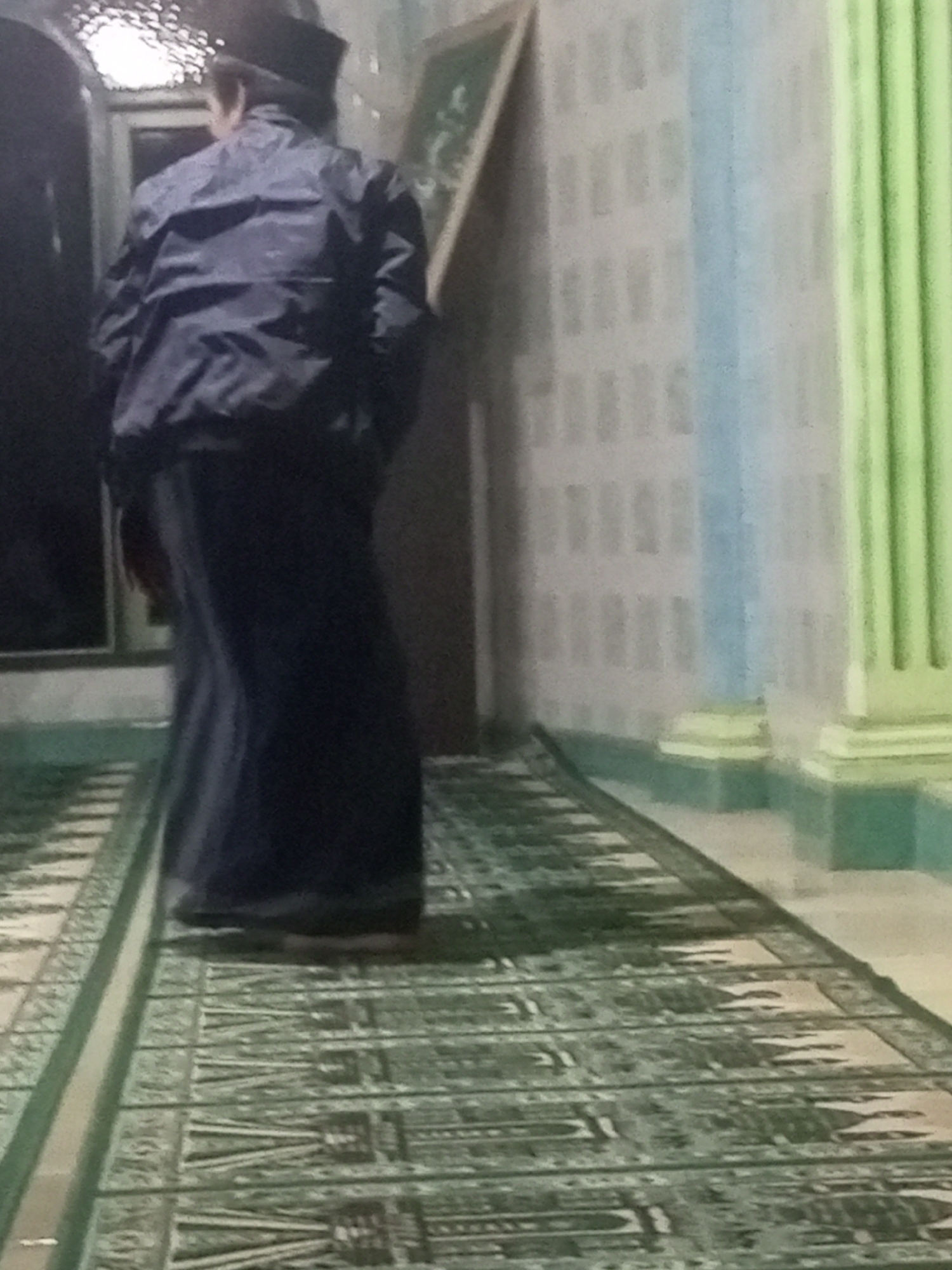The image shows a person from behind, wearing a dark jacket and a traditional cap, standing on a patterned carpet. The carpet appears to be a prayer rug, suggesting the location could be a mosque or a prayer room. The background features a wall with a pale blue and white striped pattern, a vertical green pillar, and a framed picture. The lighting is dim, and the overall scene has a slightly blurry quality. Based on the description of the carpet and the presence of a prayer rug, it is plausible that this scene is taking place in a mosque in Andongsili, Indonesia, a region known for its Muslim population and places of worship.