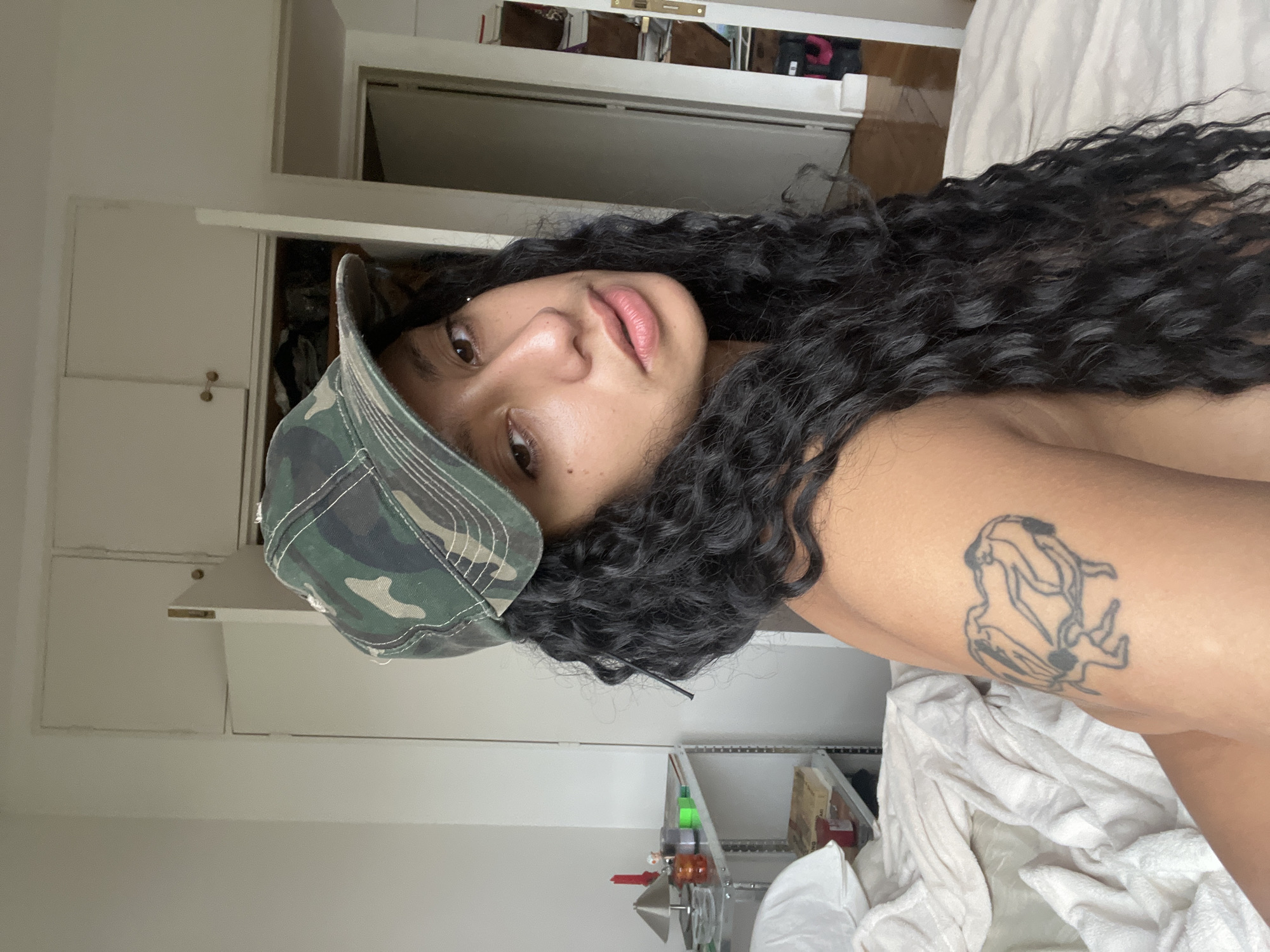 A woman with dark curly hair and a camouflage baseball cap is lying on a white sheet. She is facing the camera and has a tattoo on her right arm. The background includes a white closet and a doorway. There is no information available to indicate this photo was taken in Buenos Aires, Argentina.