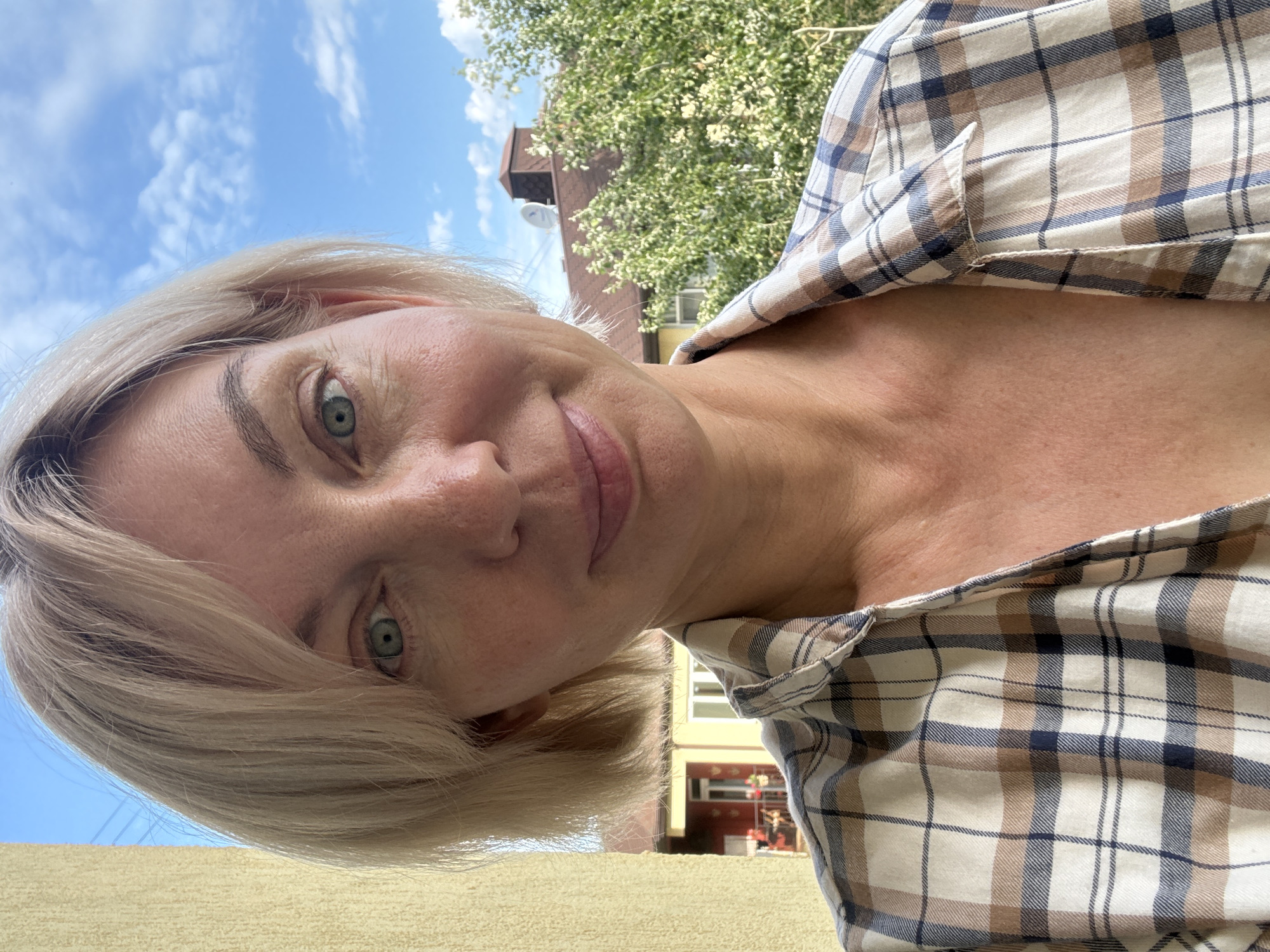A medium shot, selfie-style photograph shows a woman with blonde, chin-length hair and blue-green eyes smiling slightly. She is wearing a collared, plaid shirt in shades of beige, white, and navy blue. The background is a bright, slightly cloudy blue sky with the tops of trees and buildings visible. The image is taken outdoors. There is no indication that this image was taken in Sochi, Russia.