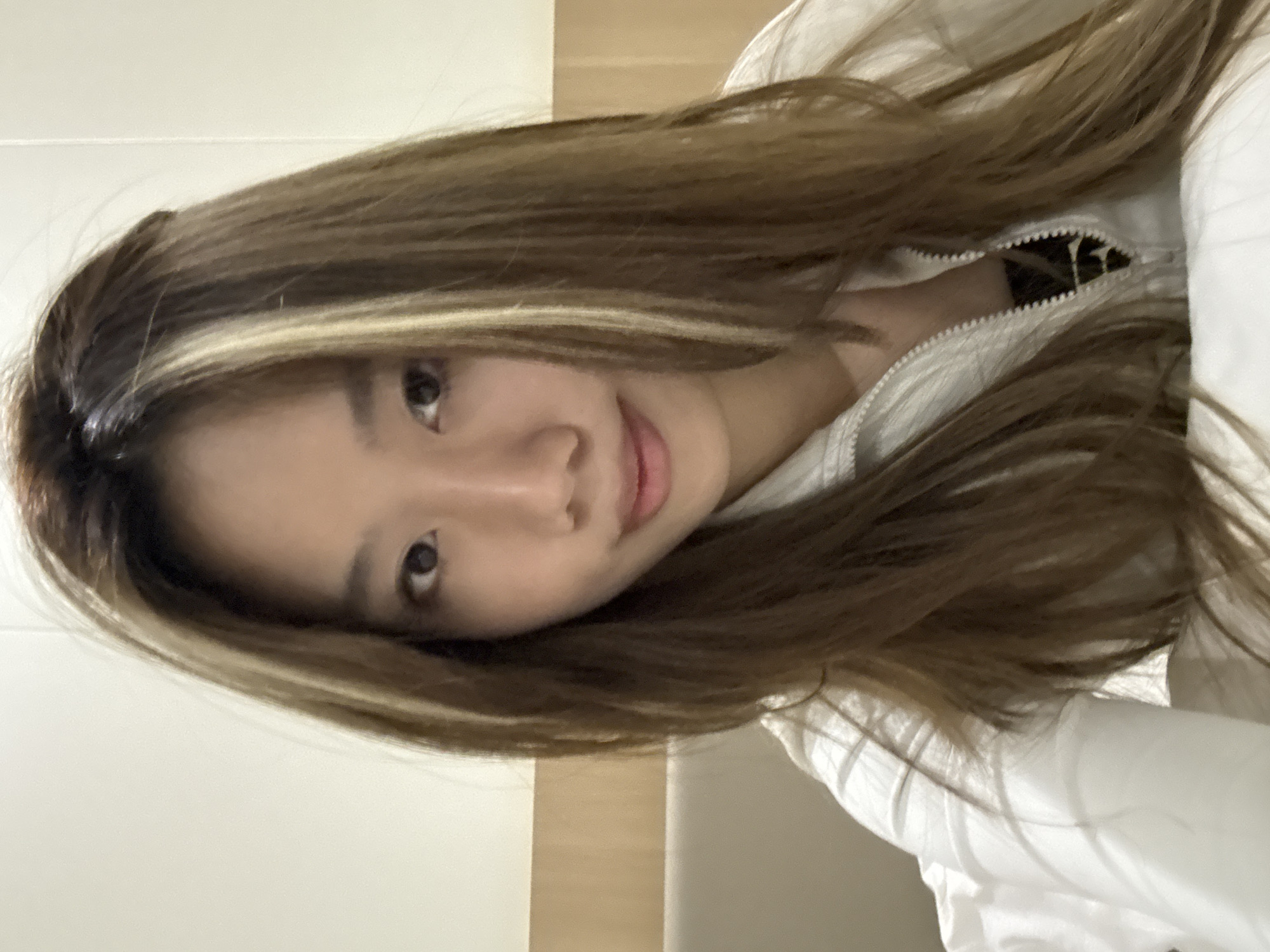 The image depicts a young Asian woman with brown hair and blonde highlights. She is wearing a white jacket. The background is a light beige wall. There is no information in the image to indicate it was taken in Dunhuang City, China.