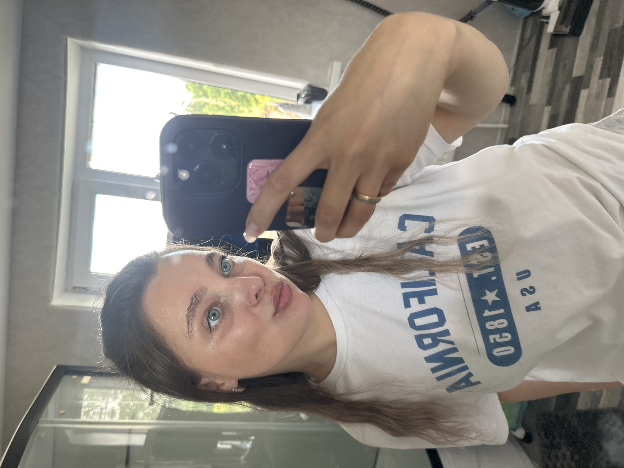 A young Caucasian woman with light brown hair and blue eyes is taking a selfie in what appears to be a bathroom. She is wearing a white t-shirt with blue lettering that reads "CALIFORNIA EST. 1920 USA". The woman is holding a black smartphone in her right hand, and her left hand is resting on her hip. Her makeup is minimal, and she is wearing a thin gold ring on her left ring finger. The background shows a window with natural light streaming in, and what looks like a cabinet or closet to the left. The floor is tiled in a wood-like pattern. The overall impression is of a casual, everyday moment. The location is not specified as being in Voznesenka, Russia.