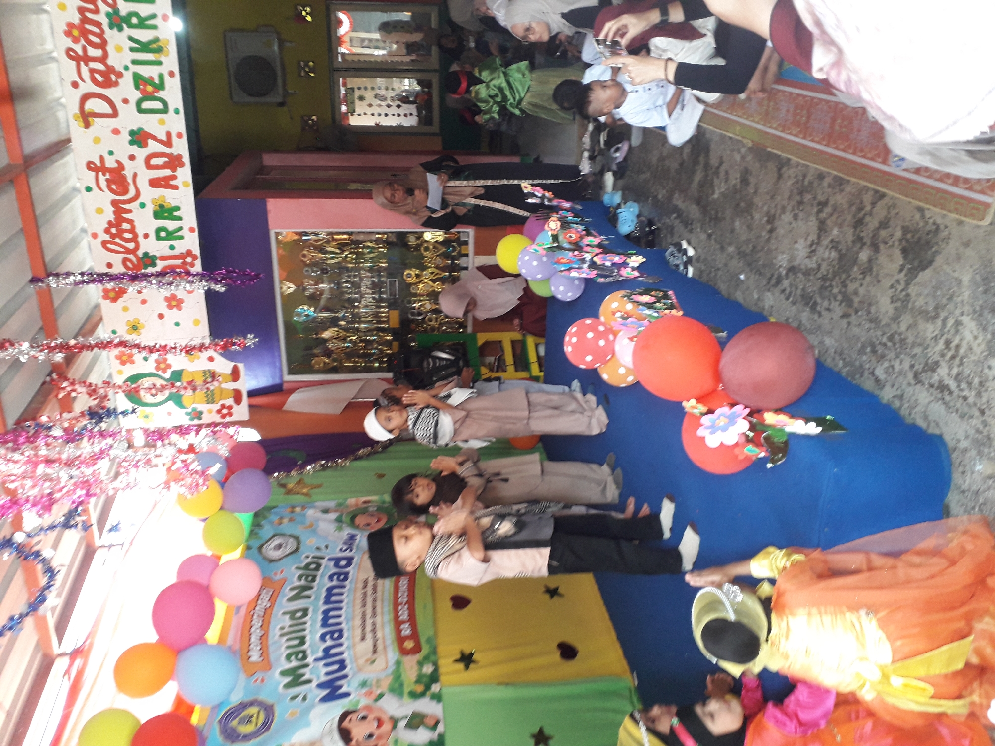 The image depicts an outdoor event, possibly a school or community gathering, taking place under a decorated roof. In the foreground, children in traditional attire are positioned on a blue stage, some with hands clasped in prayer. They are facing towards the viewer, with a banner behind them that reads "Maulid Nabi Muhammad SAW". Several balloons in various colors are visible, some clustered together.
Further back, a woman stands at a podium, addressing the audience, while other attendees, including children and adults wearing hijabs, are seated or standing around the event space. A display of trophies is visible in a glass case. The environment appears to be a covered outdoor area, with a concrete floor and possibly the side of a building or structure visible in the background. The event is adorned with colorful garlands and decorations, suggesting a festive occasion. The presence of balloons, banners, and people gathered indicates a celebration. The mention of Cankuang, Indonesia, suggests the geographical context of this event.