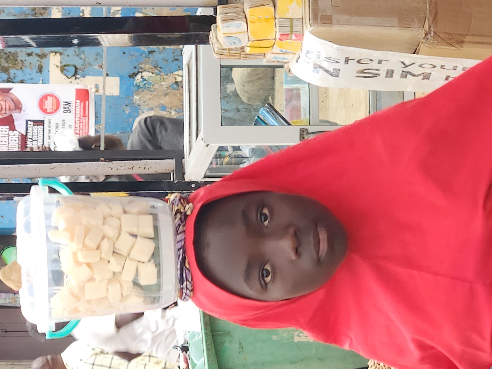 A young girl wearing a red hijab balances a clear plastic bucket filled with cubed food on her head. The bucket has light blue handles. She stands in front of a stall in Jos, Nigeria, with a green trash bin to her left and a glass-fronted display case to her right. Behind her, a blue and white weathered wall is partially covered with posters, one of which features a man and text in English, including "GREATER HONOUR" and "9AM AUDITORIUM." Stacks of what appear to be money wrapped in paper are visible on the right side of the frame, next to cardboard boxes. A white banner with black text that reads "ister Youn SIM" is attached to one of the boxes. The overall scene suggests a bustling marketplace environment in the city.