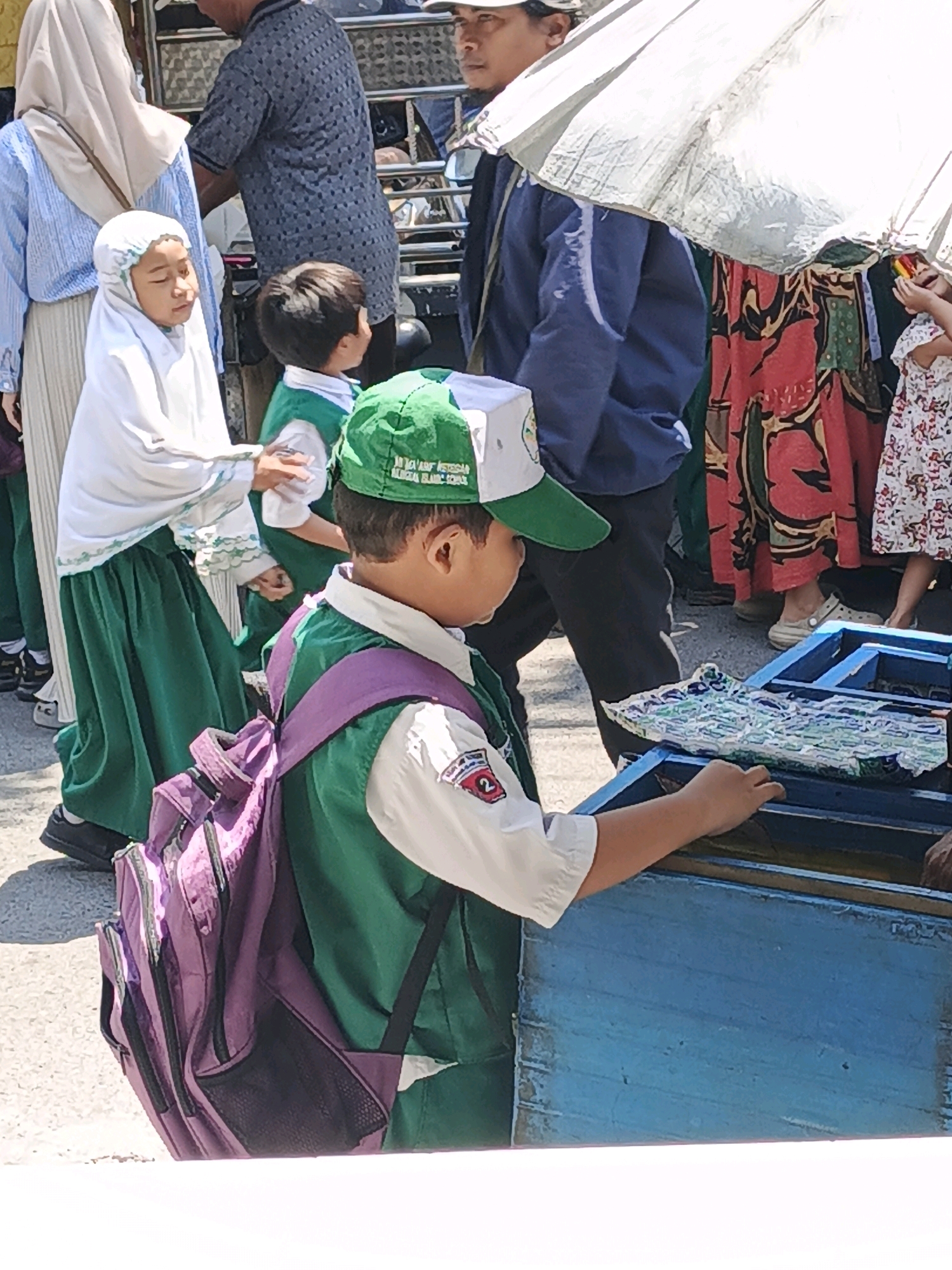 The image shows a group of people, likely students in uniform, in what appears to be a public area in Ketegan, Indonesia. In the foreground, a young boy wearing a green and white cap and a green vest over a white shirt is looking down at a blue wooden structure. He has a purple backpack on his back. To his left, a young girl in a white hijab and green skirt is looking down, and behind her, another child in a green uniform is visible. Adults are present in the background, with one person holding a large white umbrella. The scene suggests an outdoor event or activity, possibly related to a school. The ground appears to be paved, and there are other individuals in the background, some in traditional clothing. The lighting indicates it is daytime, with sunlight casting shadows.