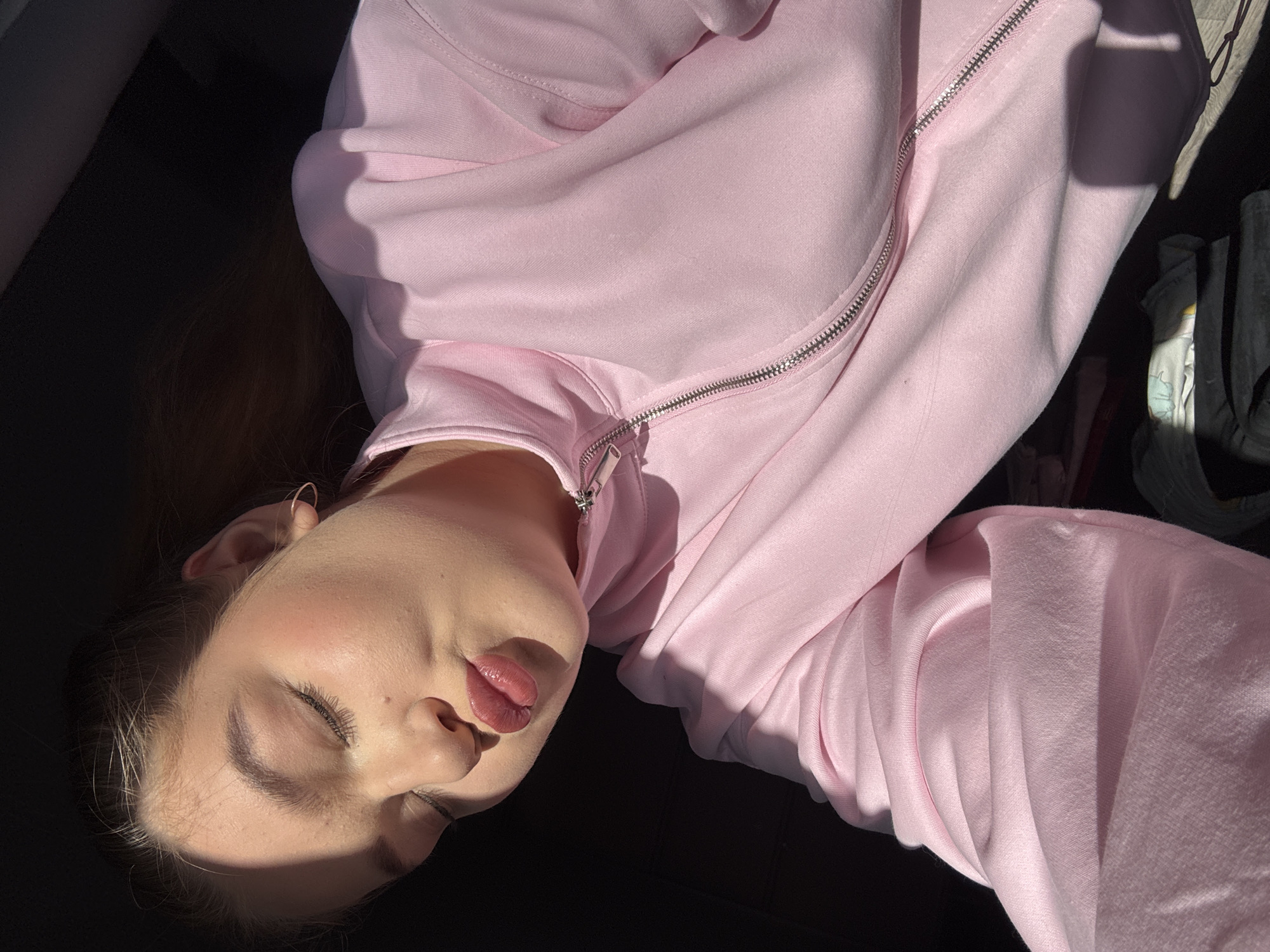 A young woman lies on a dark surface, likely a car seat, wearing a light pink zippered sweatshirt. Her eyes are closed and her lips are slightly parted. The scene is illuminated by strong sunlight, casting shadows across her face and clothing. The location, Chaikovsky, Russia, is not discernible from the image.