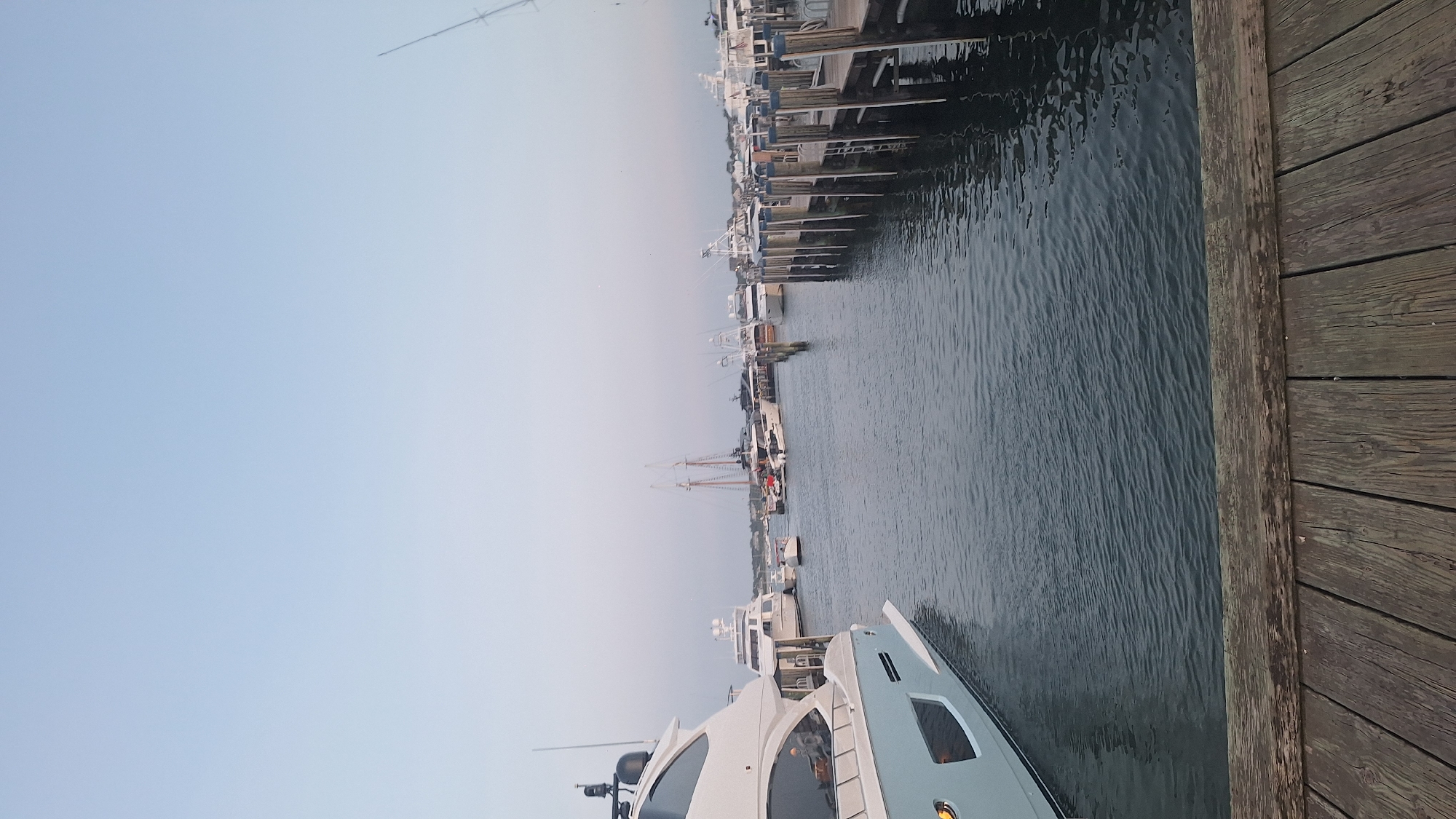 The image depicts a serene marina scene in Nantucket, United States of America, likely during twilight. The foreground is dominated by a weathered wooden dock, its planks showing signs of age and use, with a dark, rippling body of water stretching out before it. Several boats are moored in the water. On the left, a large, modern light blue and white yacht is prominently featured, its sleek design evident. Further back, a variety of other vessels are visible, including smaller motorboats and at least one classic sailing ship with tall masts. The boats are docked along wooden piers that extend into the water. In the background, a hazy shoreline with buildings can be seen, suggesting a populated area. The sky above is a pale, clear blue, transitioning to a lighter hue near the horizon, indicating the setting or rising of the sun. No people are clearly visible in the image, and the overall atmosphere is one of calm and quiet.