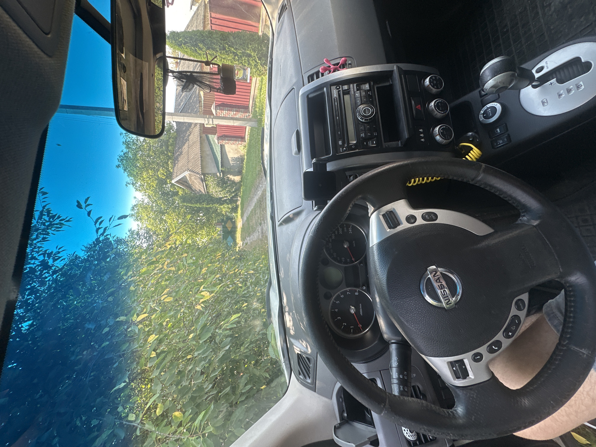 The image is taken from inside a Nissan vehicle, looking out through the windshield. The interior of the car shows a steering wheel with a Nissan logo, a dashboard with gauges, and the center console with a gear shifter and car stereo. Outside the car, a dirt road is visible, lined with lush green trees and bushes. In the background, there are houses with red roofs and fences, suggesting a residential area. The sky is a clear blue with some sunlight filtering through the trees. The photo is taken in Shatava, Ukraine.
