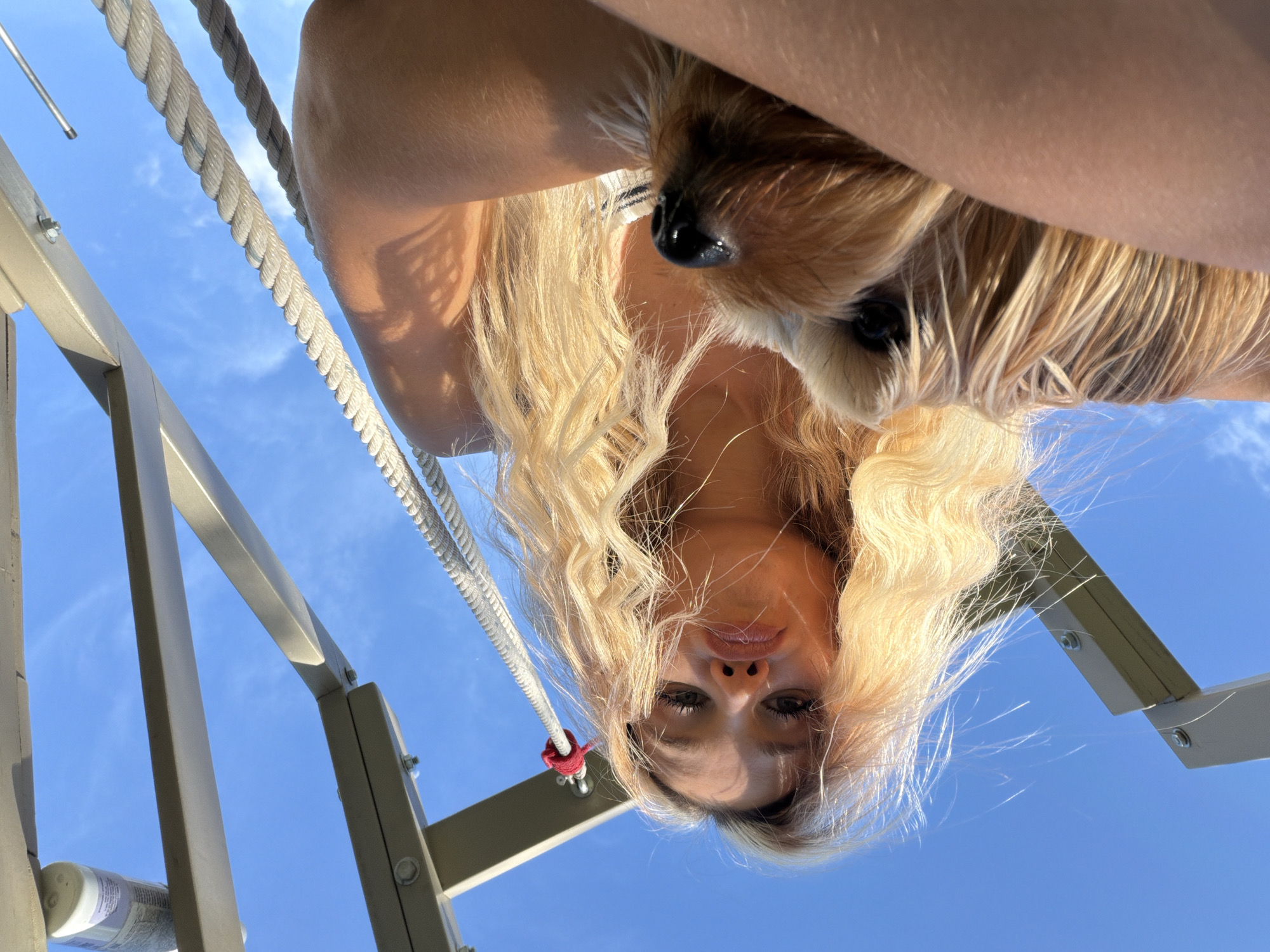 An overhead, low-angle, wide shot captures a young blonde woman holding a small, tan dog against a bright blue sky. The woman is positioned in the lower center of the frame, looking directly at the camera with her lips pursed. Her long, wavy blonde hair is windswept. The dog, which appears to be a Yorkshire Terrier, is held in her arms, close to her face. The background consists of a clear blue sky with some wispy clouds and the metallic structure of what appears to be a playground or swing set, with thick ropes hanging down. The image was taken outdoors on a sunny day in Thep Krasatti, Thailand.