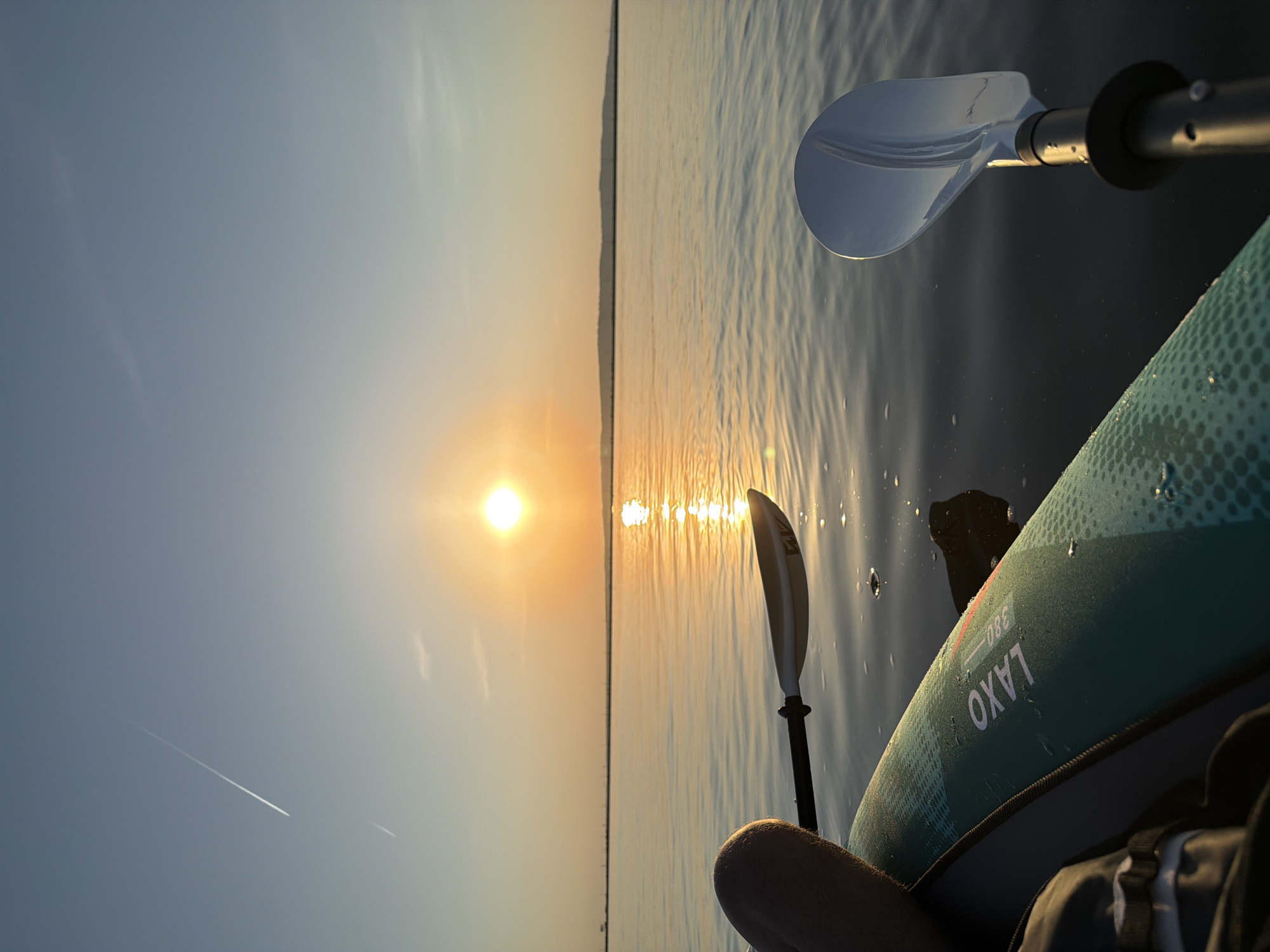 The image shows a view from a kayak or paddleboard on a body of water, likely a lake or wide river, at sunset. The sun is positioned in the center of the frame, reflecting brightly on the water surface. A shoreline is visible in the distance, appearing as a dark line against the sky. The foreground includes parts of a paddle and the edge of the kayak or paddleboard, marked with "LAXO 380". The sky is clear with the sun. Given the prompt, it is suggested that this image was taken in Chaikovsky, Russia. There are no people fully visible, but a leg is visible in the lower right.