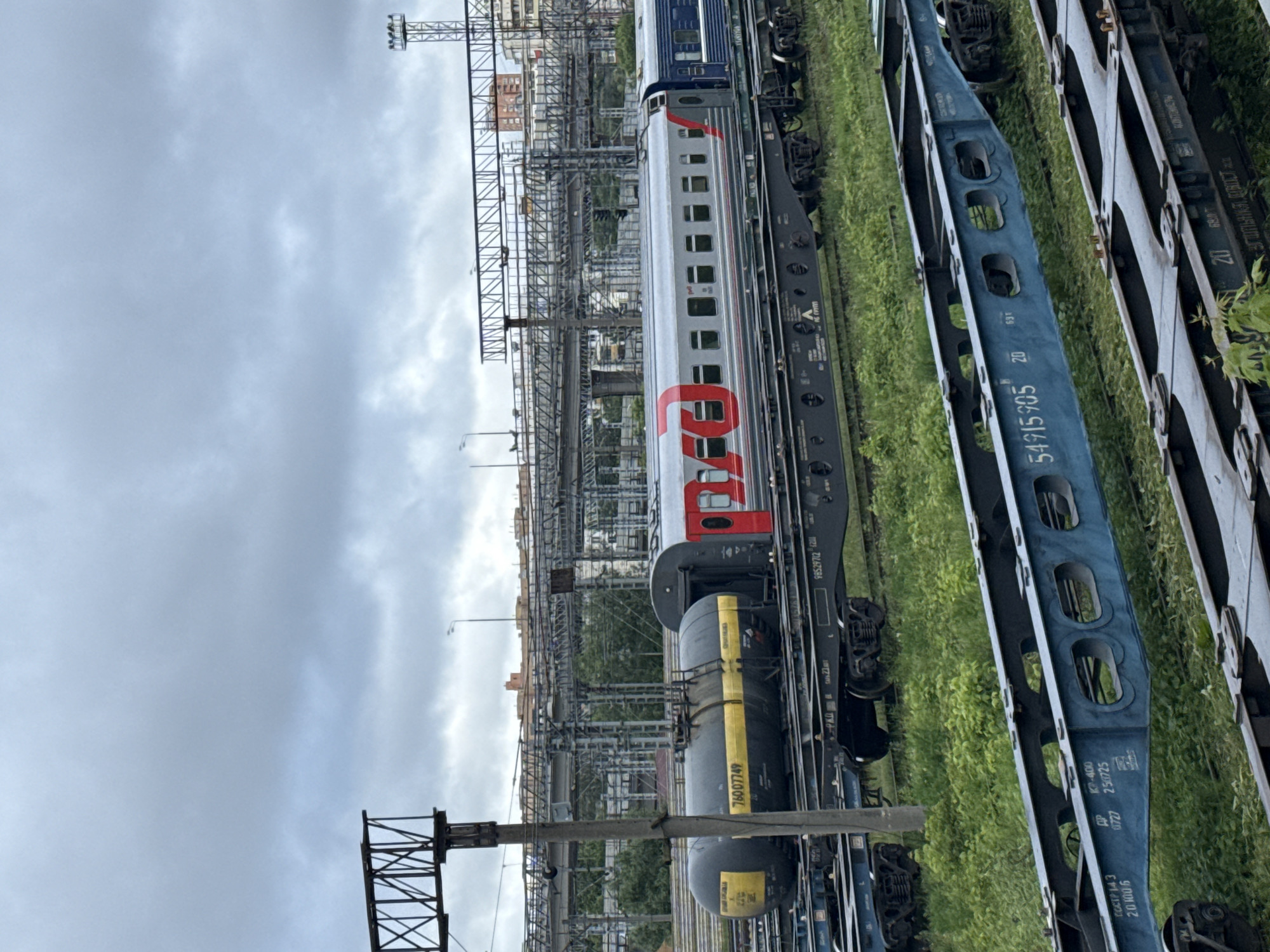 The image shows a train yard in what appears to be Moscow, Russia, given the "РЖД" logo on the train. The scene includes railroad tracks, train cars (tanker, passenger, and blue flatcars). An overhead catenary system is visible. Buildings are in the background under a cloudy sky.