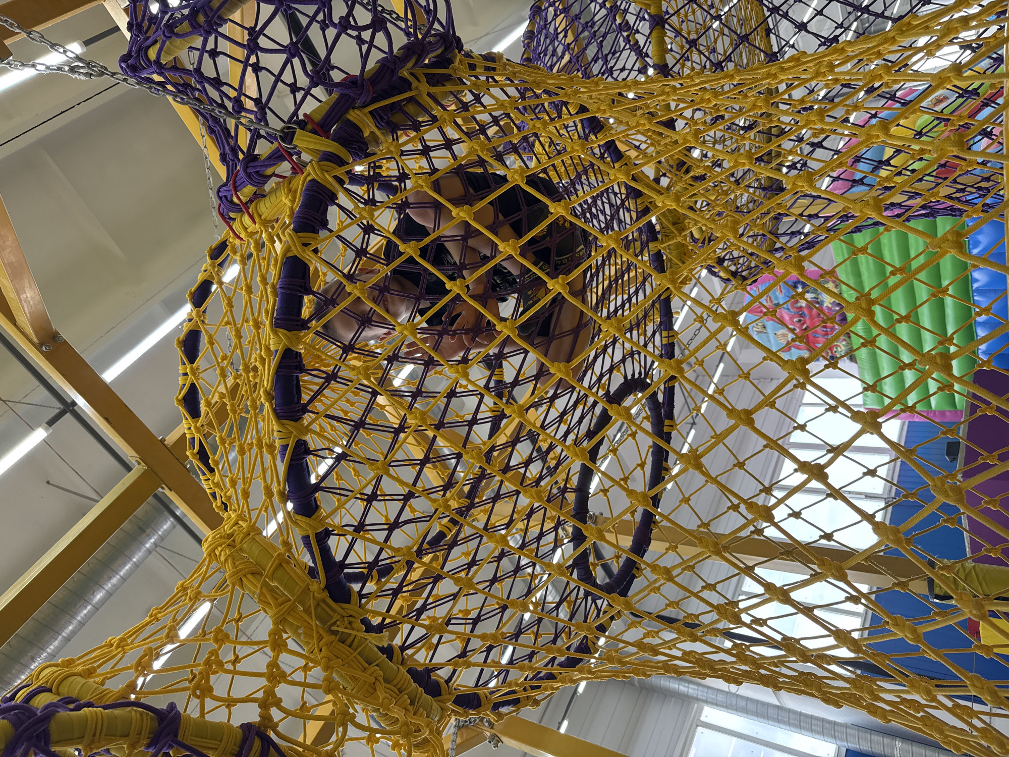 A young boy is sitting inside a large net structure in an indoor playground. The net is made of yellow and purple ropes, creating a diamond pattern. The boy is wearing a black t-shirt and dark shorts. He is looking towards the camera with a slight smile and has his hands on the ropes. The background shows parts of the playground, including brightly colored inflatable structures, and the ceiling of the building with fluorescent lights and yellow beams. The location is the "Chaikovsky Adventure Park" in Chaikovsky, Russia.