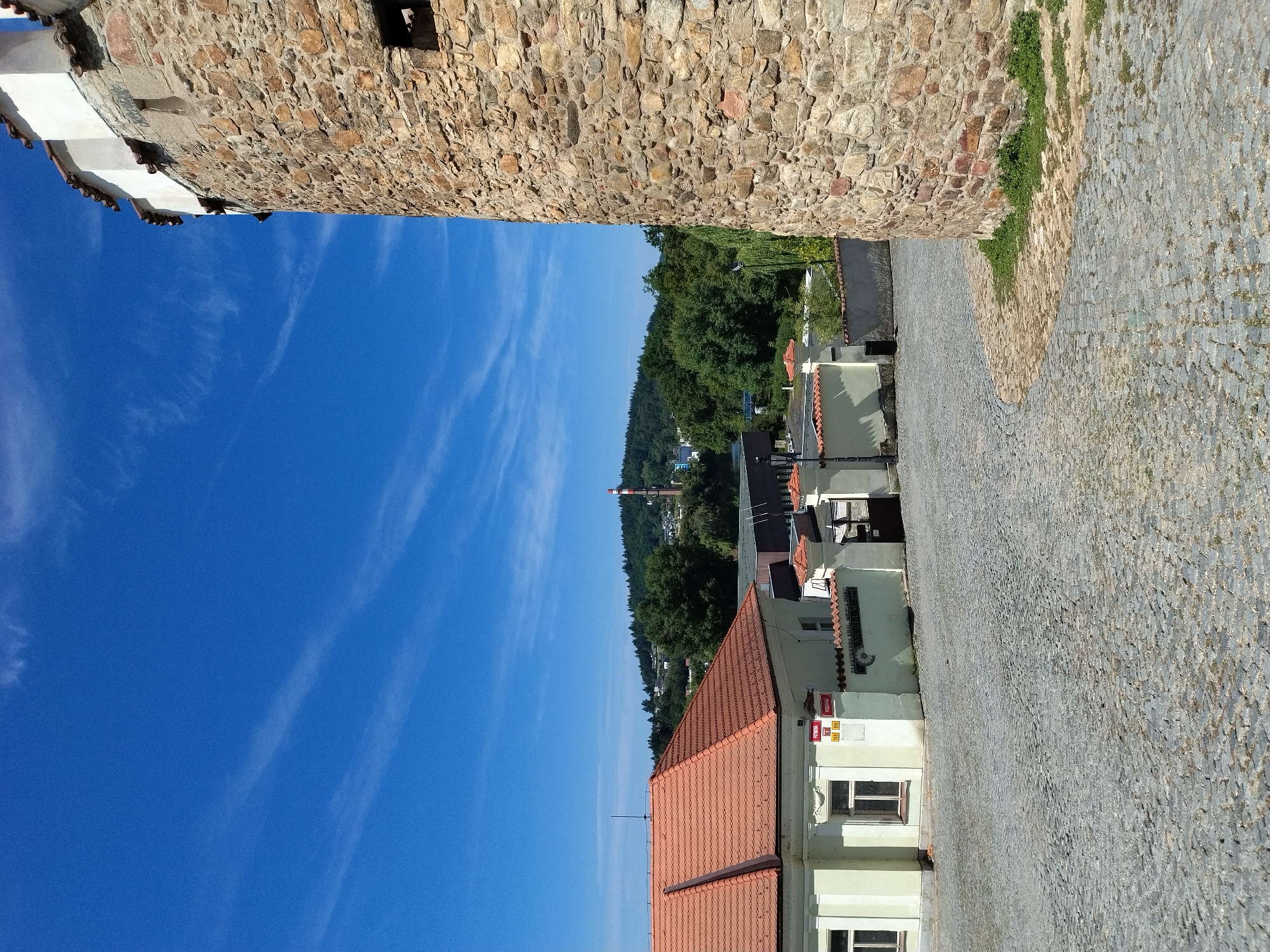 The image depicts a view of the city of Písek, Czechia, with a prominent stone tower on the right side, suggesting it is part of a historical fortification. In the foreground, a cobblestone street curves into the scene, leading towards a light-colored building with a red-tiled roof. Behind this building, a low wall with red-tiled coping separates it from what appears to be a public area or courtyard, featuring an ornate gate. Further in the background, a hill covered in trees is visible, and on its crest, a tall industrial chimney with red and white bands stands out against the bright blue sky, which is streaked with wispy white clouds. The overall scene conveys a sunny day in a European city, blending historical architecture with industrial elements and natural landscape.
