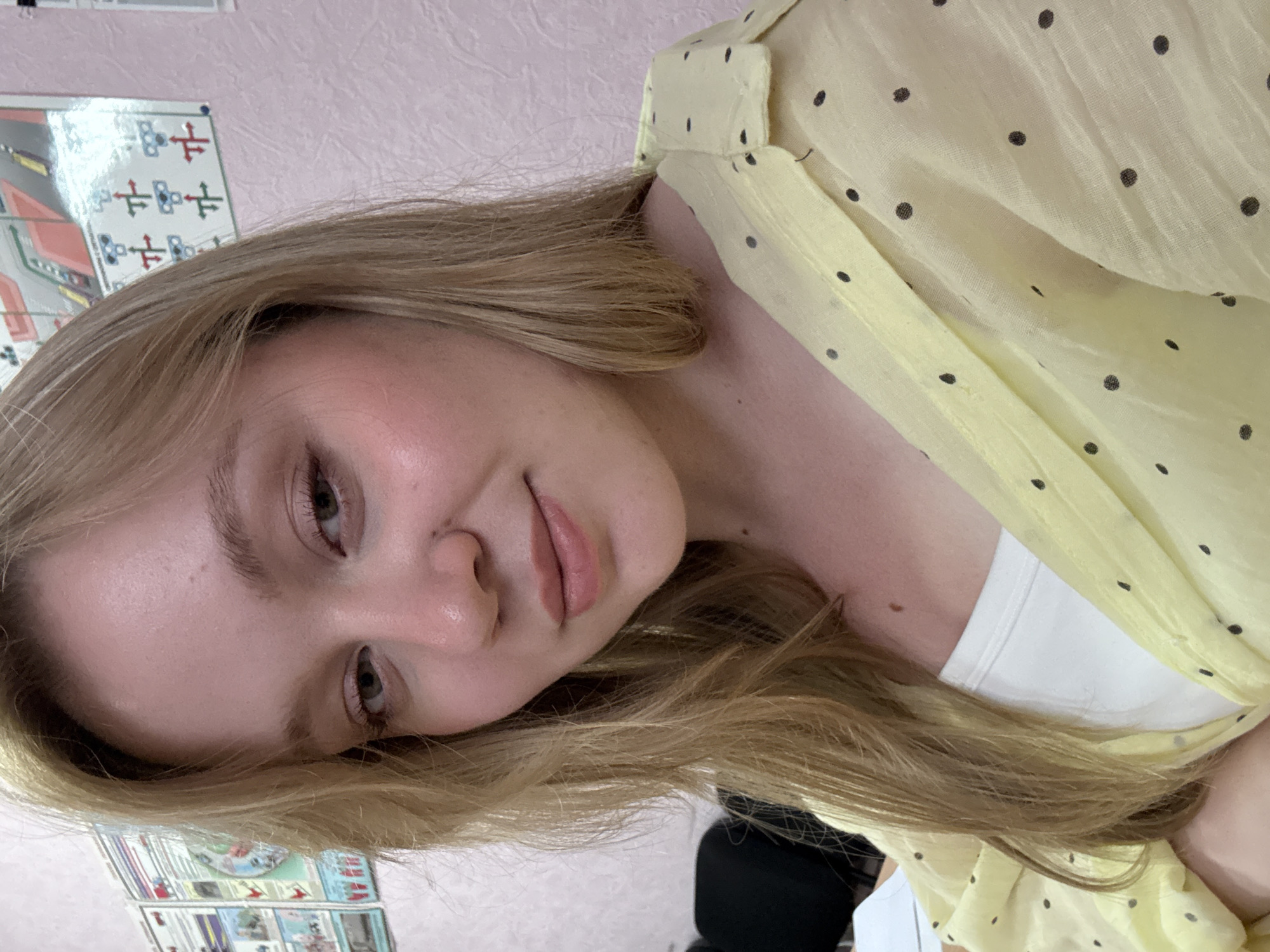The image is a close-up, chest-up, selfie-style photograph of a young Caucasian woman with shoulder-length blonde hair. She is indoors, in what appears to be an office or classroom setting, indicated by posters on the wall behind her. The posters are colorful and appear to be educational or informational charts. The woman is wearing a light yellow, polka-dot collared shirt over a white tank top. Her skin is fair with some natural blush. She has light-colored eyes and is looking towards the camera with a slight smile. The background wall is pink with a textured pattern. There are no discernible objects in the foreground other than the woman's clothing. The lighting is natural and appears to be coming from the front. The image does not contain any specific visual cues that would place it in Chaikovsky, Russia.