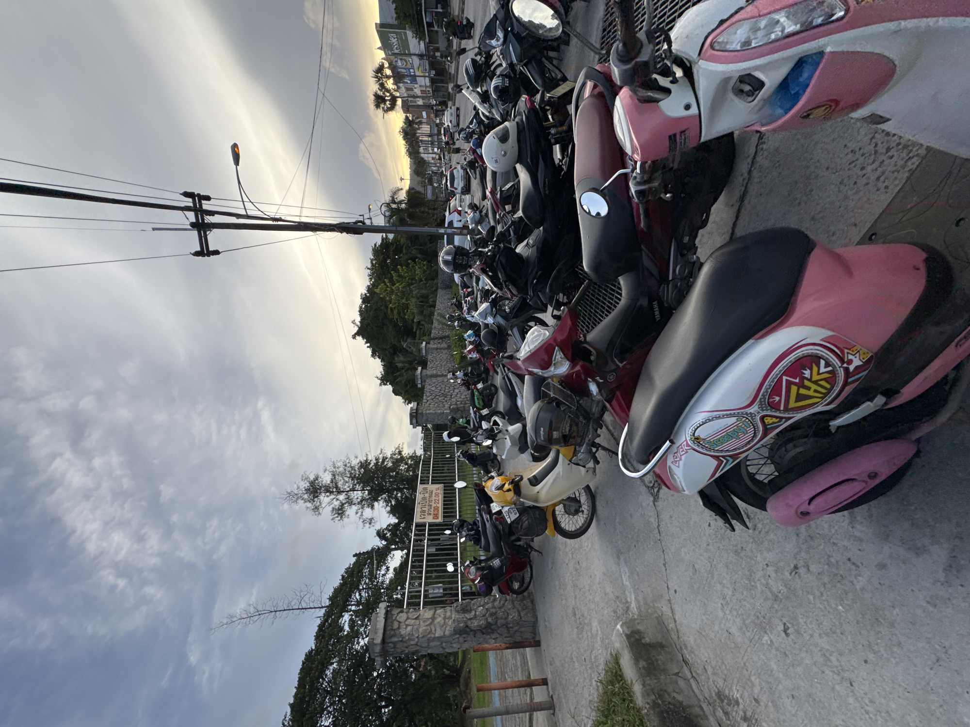 This media file depicts a row of parked motorcycles and scooters on a concrete surface, likely a parking lot or street. The scene is set outdoors, with a backdrop of trees and some buildings visible in the distance. The sky is overcast with streaks of light suggesting either sunrise or sunset. The time of day appears to be late afternoon or early evening.
The subjects are primarily the vehicles, which are predominantly scooters, with a few motorcycles interspersed. Many of the scooters are brightly colored, with prominent examples being pink and white, and yellow. Several of the scooters have helmets resting on them or attached. There are no people visible in the frame, but the presence of the vehicles suggests they are nearby.
Notable details include a street lamp visible in the distance, and a metal fence with a stone base to the left. A sign with Thai text is affixed to the fence. The overall atmosphere is one of a typical urban or semi-urban scene in Southeast Asia, likely Thailand, given the location context of Phuket City Municipality. The weather appears to be mild, with no strong indicators of rain or extreme heat. The lighting suggests a diffused light, common during twilight hours. The vehicles are parked in a somewhat orderly fashion, indicating a designated parking area.
