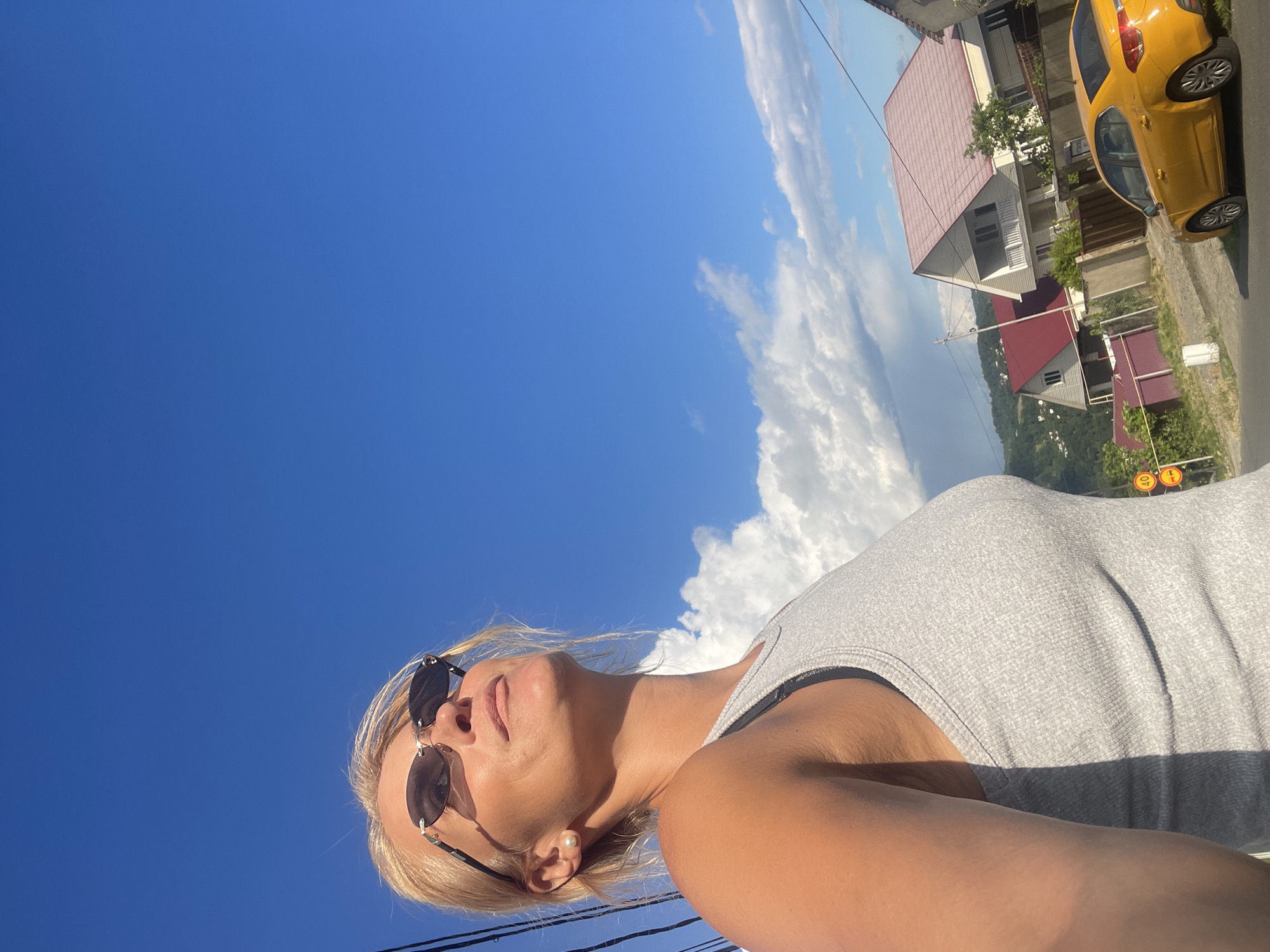 In this outdoor medium shot, a blonde woman with sunglasses and a pearl earring is captured from a low angle, looking upwards towards a vibrant blue sky dotted with fluffy white clouds. She is wearing a grey tank top. To her right, a street lined with houses and a yellow car is visible. The scene appears to be in a hilly area, with lush green trees and more houses visible in the distance. The sunlight suggests it is a bright, clear day. The image is oriented vertically. No visible text is present in the image.
