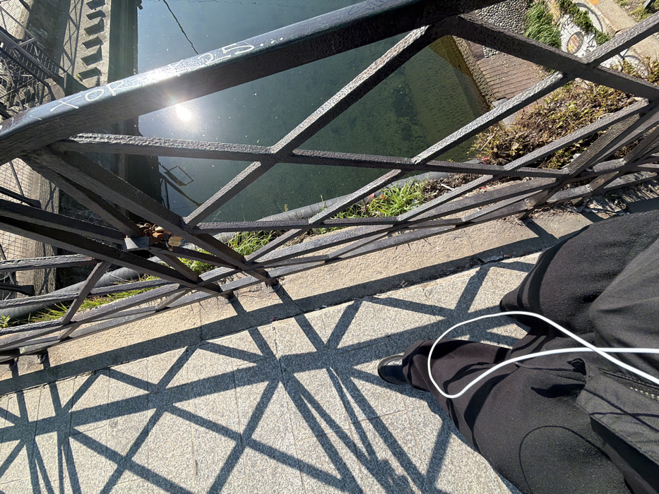 The image presents a downward, first-person perspective of an outdoor scene, identifiable as a canal-side walkway in the Navigli district of Milan, Italy. In the lower-left foreground, the lower body of an individual is visible, dressed in dark trousers and a dark outer garment, with a black shoe on the left foot. A white wired earbud cable descends over the left leg. The individual stands on a light-colored, paved surface, marked by distinct geometric shadows cast by a dark wrought-iron railing. This railing, featuring a grid pattern of horizontal and diagonal bars, extends across the mid-ground, separating the walkway from a body of water. Several small, golden-hued padlocks are attached to a horizontal bar of the railing, and white graffiti is present on one of its vertical elements. The water below the railing is a greenish canal, reflecting a bright sun flare in its upper right quadrant. Green foliage and dry vegetation are visible along the canal bank beneath the railing. Submerged objects, possibly steps or a ladder, are discernible in the water on the far side, adjacent to a textured stone wall.