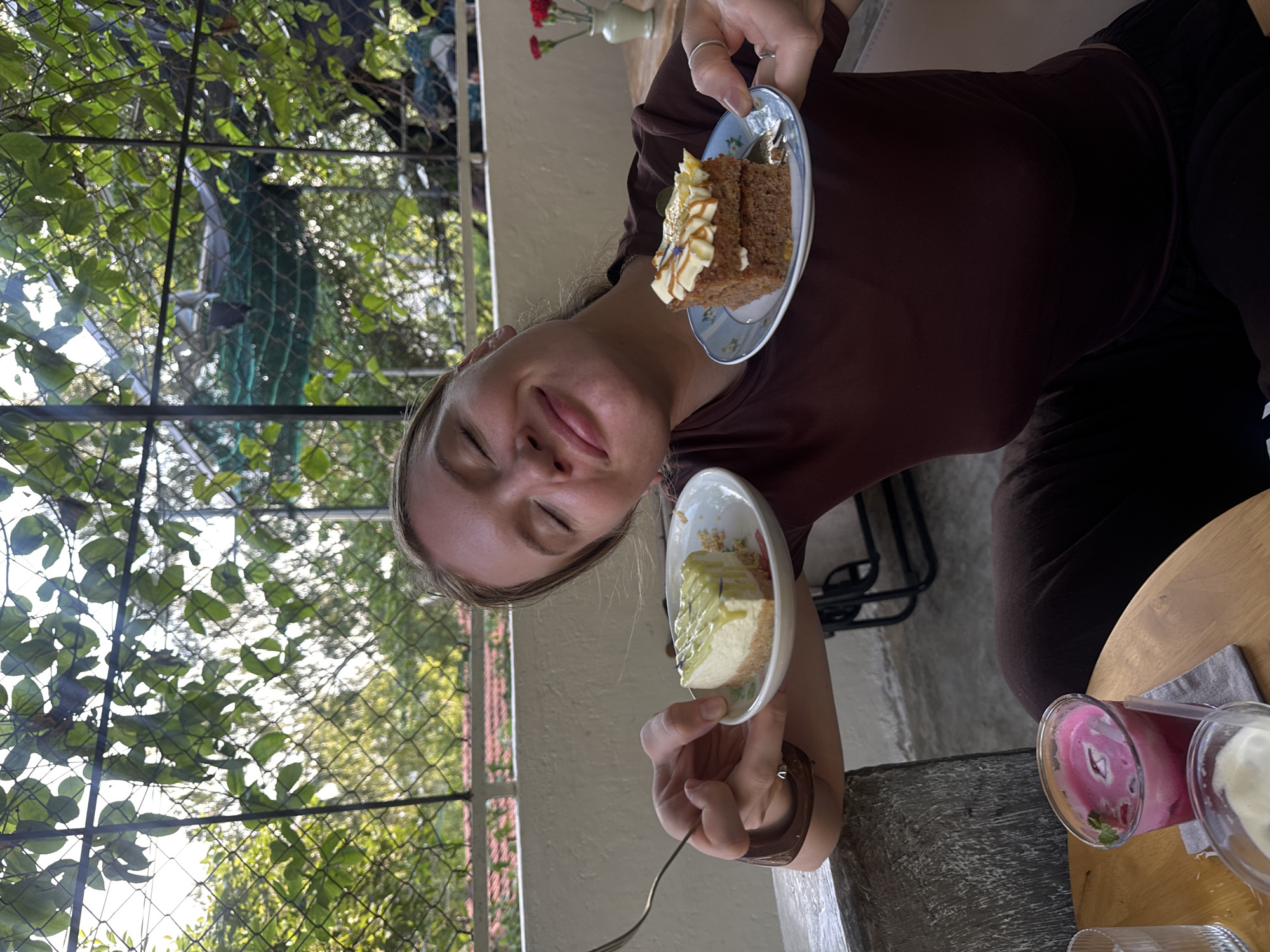 A young Caucasian woman with blonde hair tied back is sitting at an outdoor cafe, holding two small plates of dessert. One plate has a slice of cheesecake with a green topping and a crumbly crust. The other plate holds two stacked slices of cake with white frosting and a caramel drizzle, topped with sesame seeds and what appears to be edible gold flakes. The woman is smiling with her eyes closed, leaning back slightly as she presents the desserts. She is wearing a dark maroon t-shirt and dark pants. In the foreground, on a wooden table, are two glasses of drinks: one with a pinkish-purple liquid, ice, and mint leaves, and another with a white creamy substance and a straw. The cafe appears to be located in a leafy, green environment, with vines and foliage visible in the background, and a fence or netting structure. The wall behind her is a light, textured beige. There are also glimpses of other patrons and tables in the background, as well as a vase with red flowers. The overall atmosphere is casual and relaxed, suggesting a pleasant day at a cafe in Bac Nha Trang Ward, Vietnam.