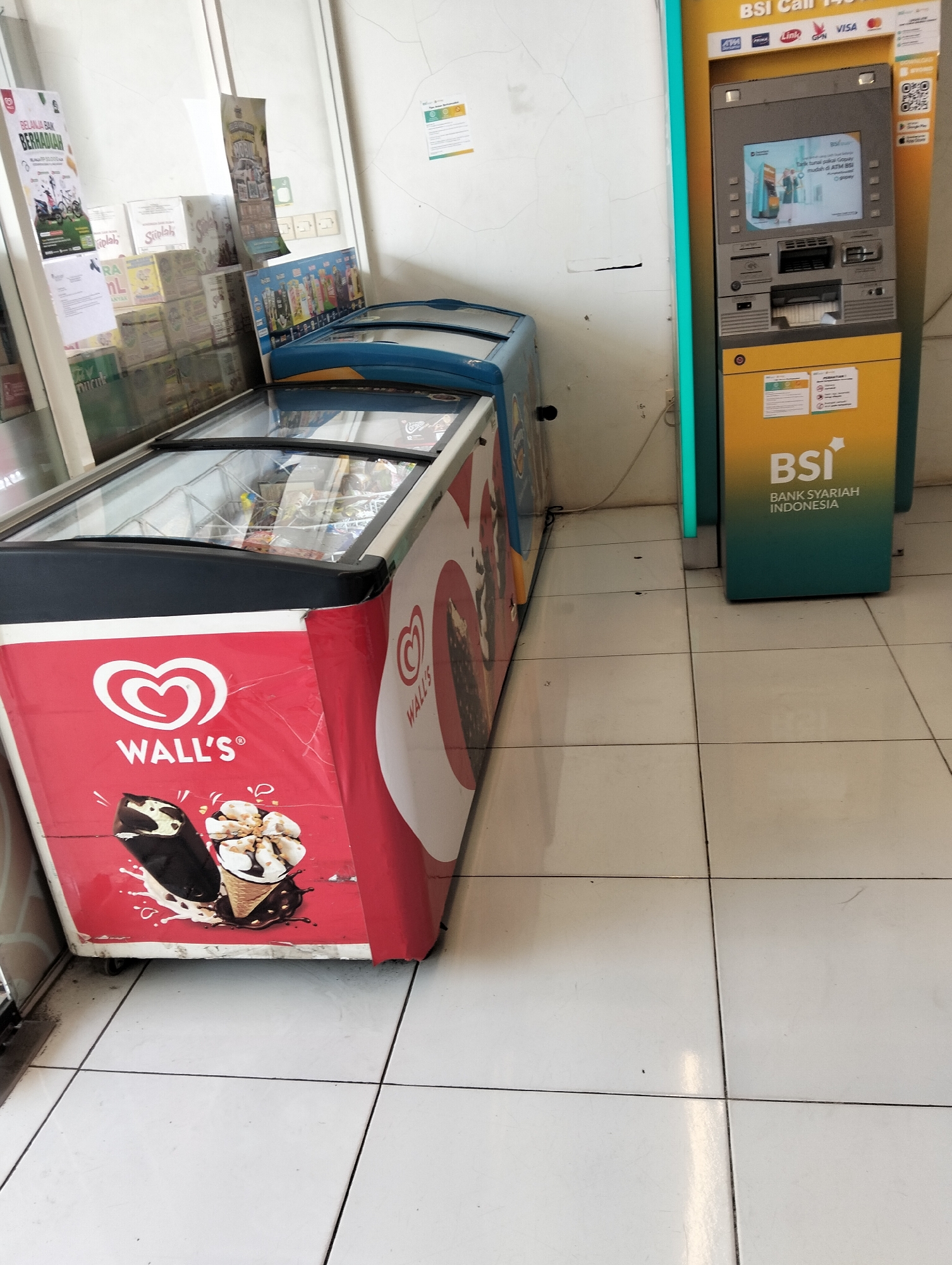In Muncar, Indonesia, a scene unfolds inside a store featuring retail displays and a banking kiosk. Dominating the foreground is a red and white Wall's ice cream freezer, adorned with images of ice cream cones and a Wall's logo. Next to it, a blue-topped freezer is partially visible. To the right, a bright yellow Bank Syariah Indonesia (BSI) ATM machine stands, its screen displaying a promotional image and various card logos such as Visa and Link. Various signs and posters are affixed to the walls and the ATM. The floor is tiled with white squares. The environment appears to be an indoor retail space, likely a minimarket or convenience store, given the presence of both ice cream freezers and an ATM. The lighting suggests it is daytime, with artificial light from within the store illuminating the scene. No individuals are present in the immediate frame.