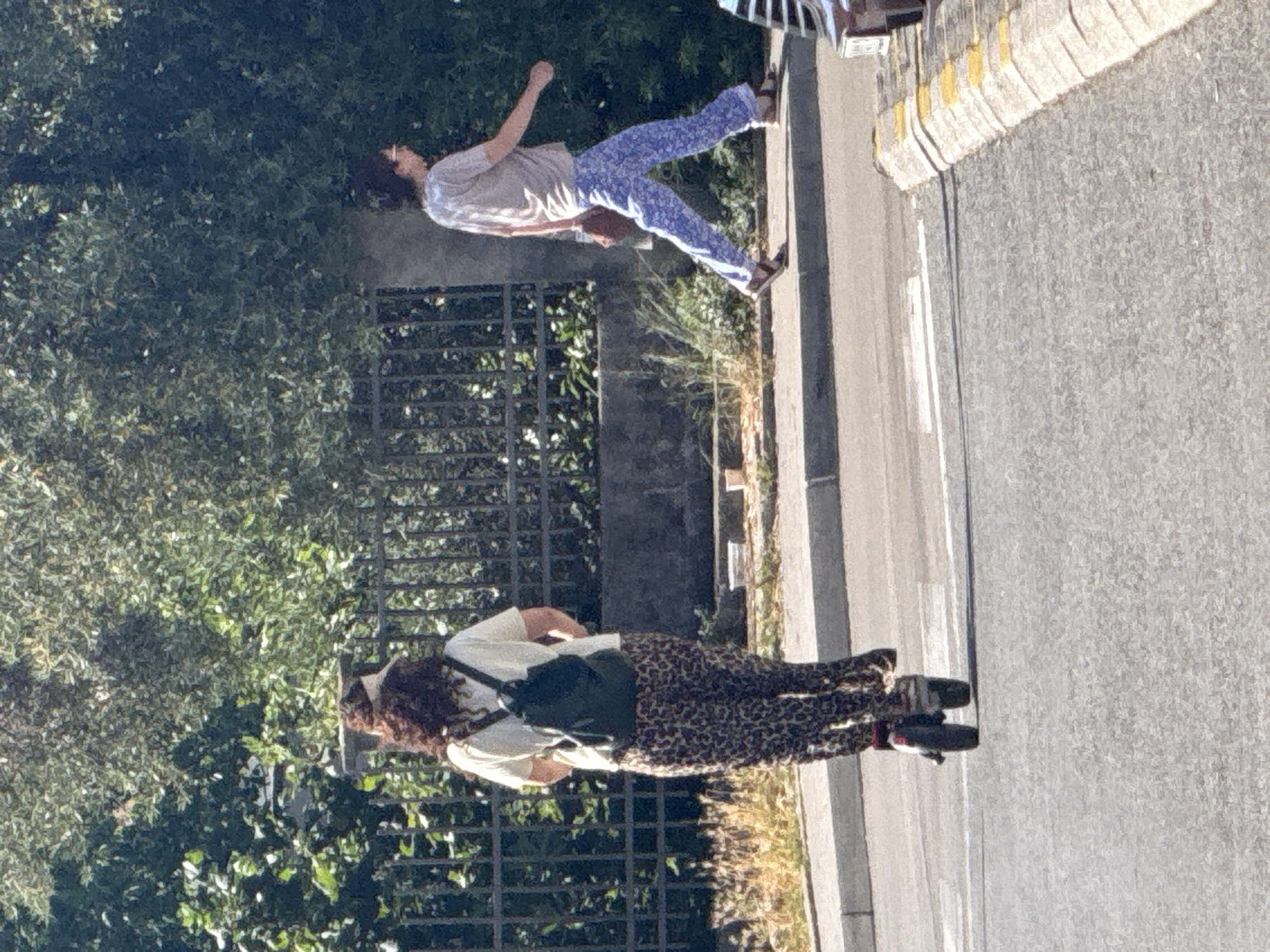 In Donji Pobori, Montenegro, a person on an electric scooter with curly brown hair and a white t-shirt, wearing leopard print pants and a dark backpack, is moving away from the camera on a paved road. To the right, a second person wearing a light-colored t-shirt and blue patterned pants is walking on a sidewalk. The background consists of green foliage and a black metal fence. The scene is illuminated by sunlight.