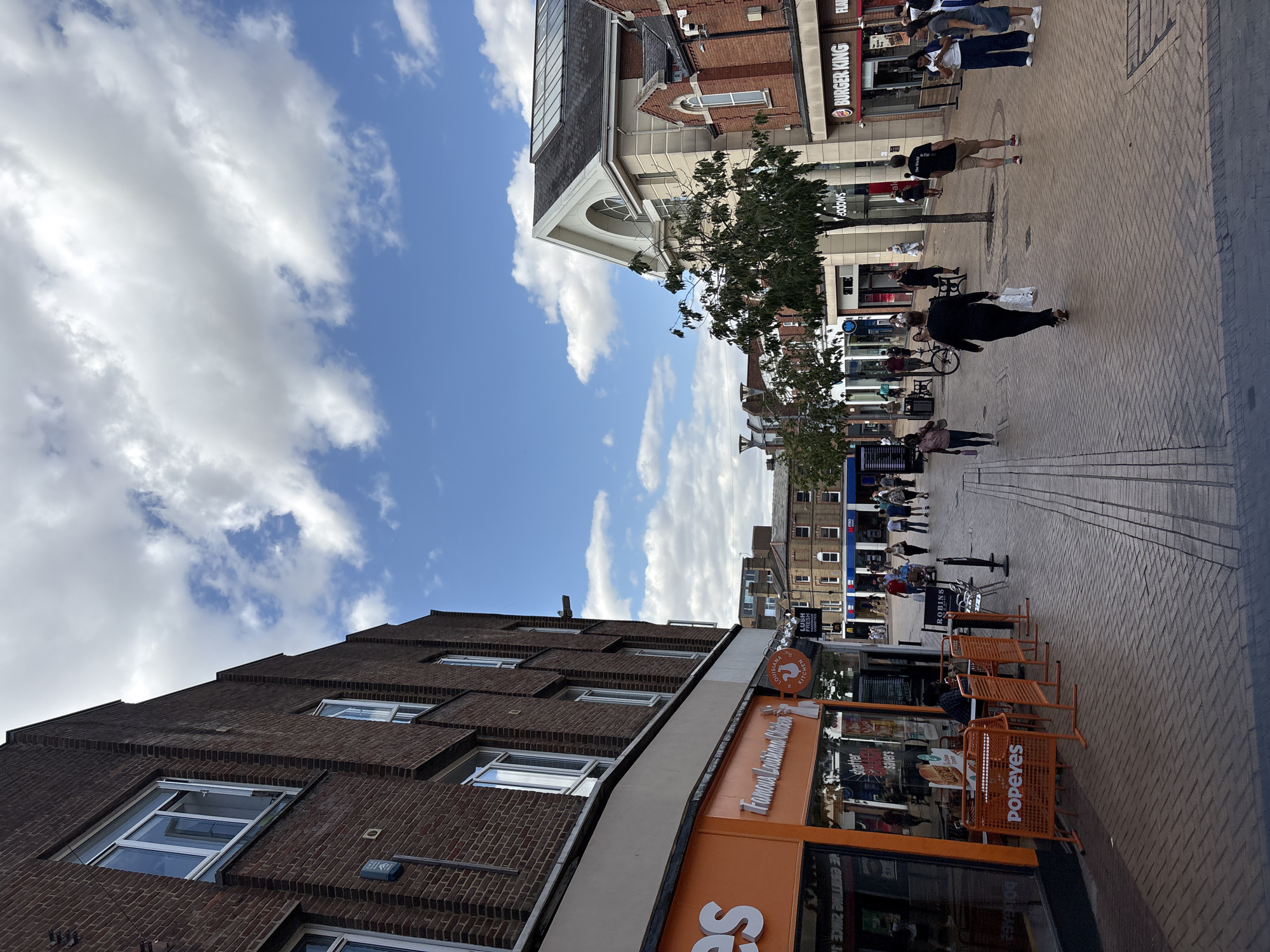 The image depicts a sunny day in Chelmsford, United Kingdom, showing a pedestrianized shopping street. On the left, a Popeyes restaurant with its signature orange signage is prominently featured. Beside it, a Burger King restaurant is visible. Several people are walking along the street, and some are seated at tables outside Popeyes. The architecture suggests a modern commercial district with a mix of brick and lighter-colored buildings. The sky is blue with scattered white clouds. The street is paved with bricks, and there are trees lining the walkway. The overall scene is one of a typical urban environment with commercial activity.