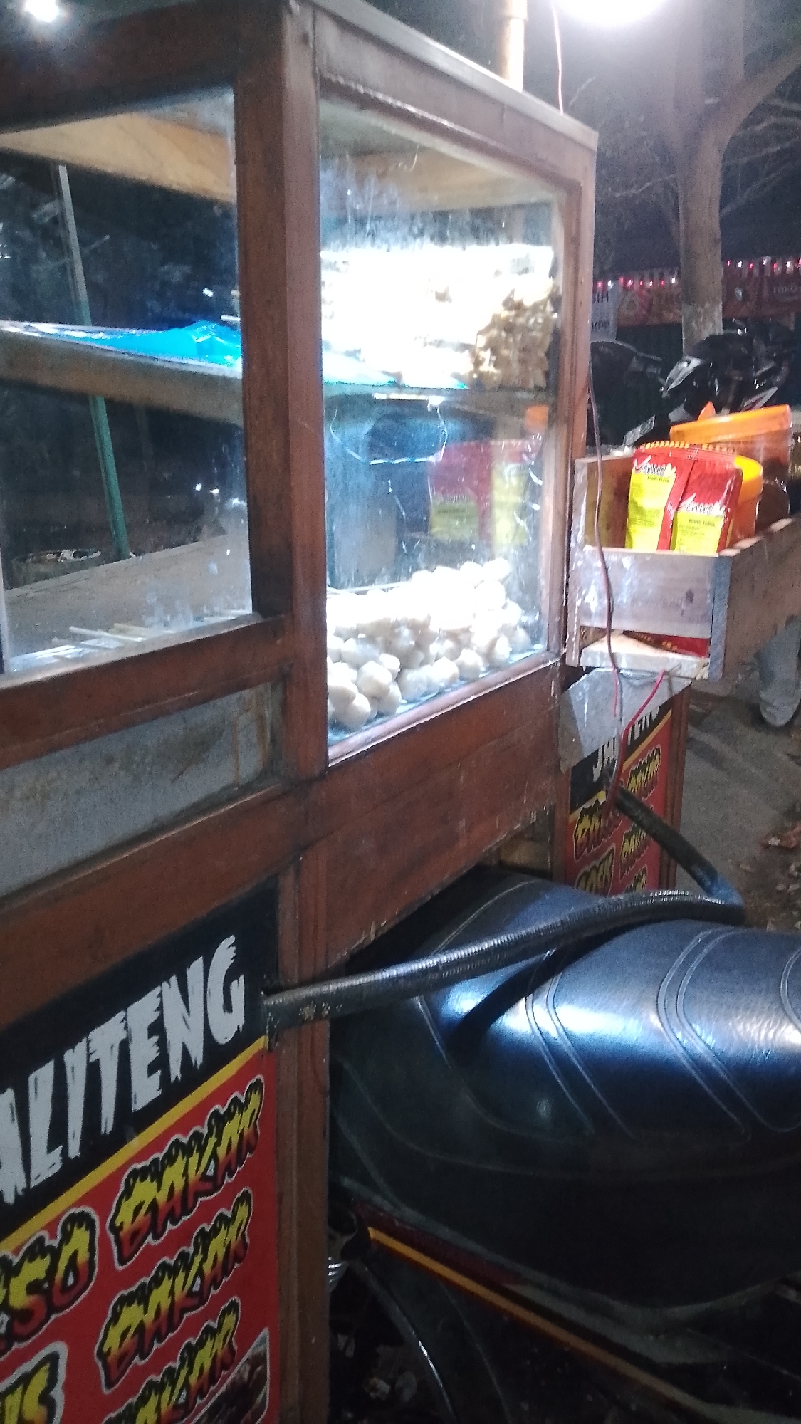 The image shows a street food cart, likely in the city of Karanglangu, Indonesia. The cart has a wooden frame and glass display cases. Inside the cases, there are many small, round, white food items, possibly meatballs or dumplings, displayed on skewers. Several packets of food items, possibly snacks or seasoning, are visible on a shelf to the right of the display cases. A motorcycle seat is visible in the foreground, partially obscuring the lower part of the cart. The cart has signage with Indonesian text, including "ALITENG" and "SOSO BAKAR". The overall lighting suggests it is evening or night.