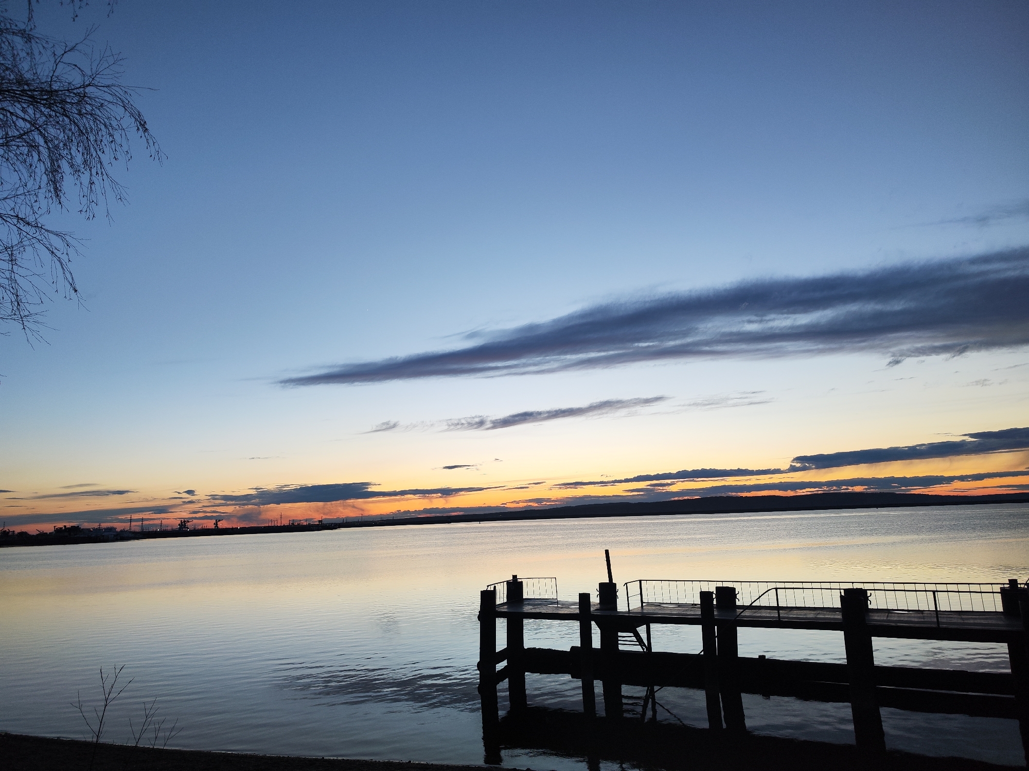 The image depicts a pier extending into a body of water, likely a lake or large river, at sunset. The sky is a gradient of blue transitioning to orange and yellow near the horizon, with scattered clouds. The pier is silhouetted against the water and sky, appearing dark. The water reflects the colors of the sky. In the background, across the water, is a faint skyline that appears to be the city of Chaikovsky, Russia, with industrial structures and a suggestion of smoke or steam rising. Some bare tree branches are visible in the upper left corner. The photograph has a tranquil, serene quality, focusing on the natural beauty of the sunset and the stillness of the water.