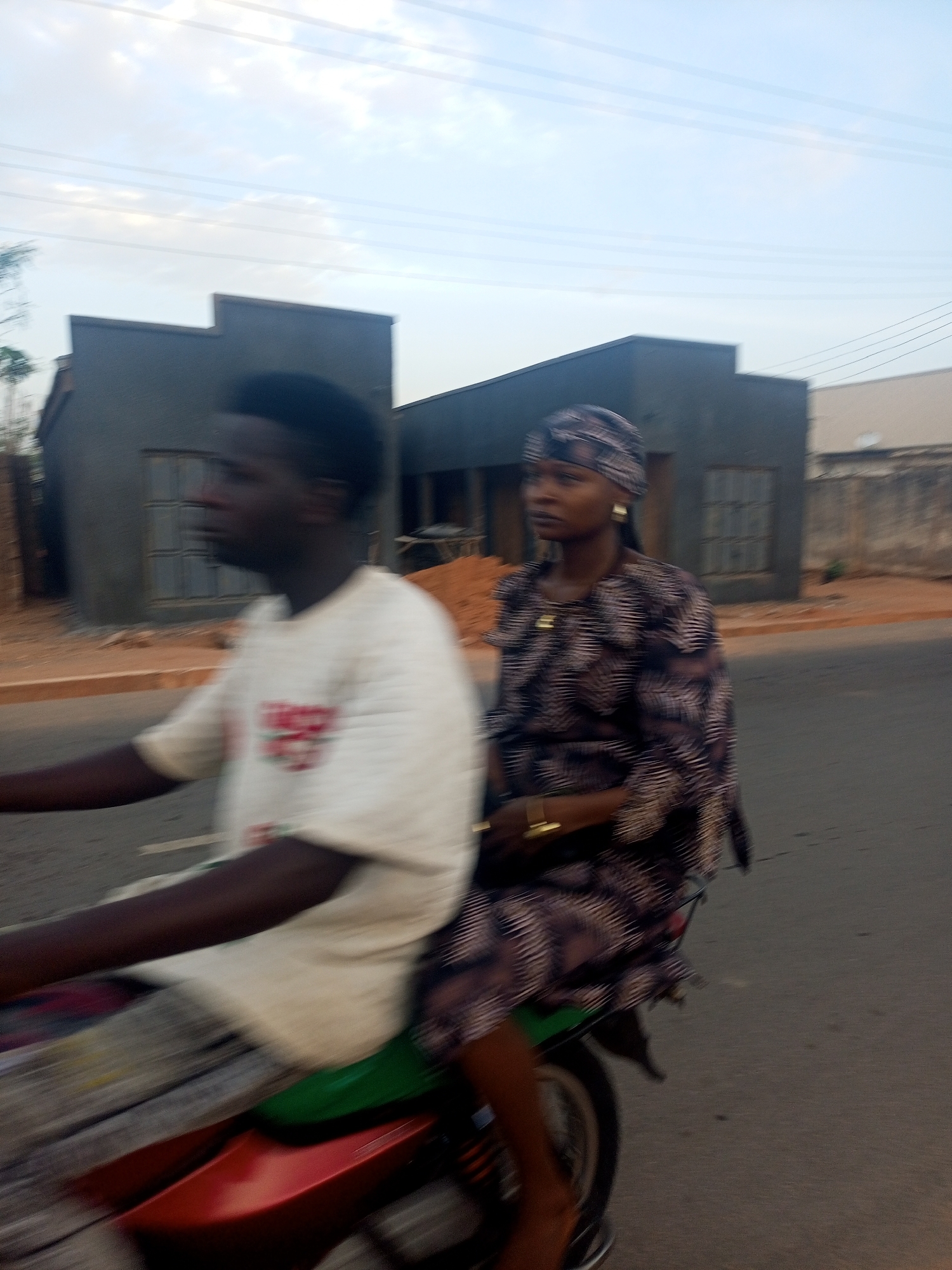 A man and woman are riding a red motorcycle on a street in an urban area. The man, who is driving, is wearing a white t-shirt and patterned pants. The woman is wearing a dark patterned outfit with a head covering. In the background are unfinished grey buildings with window frames. The street is paved and there is a partial view of a fence in the distance. Overhead are visible power lines. The city is identified as Unknown, Unknown.