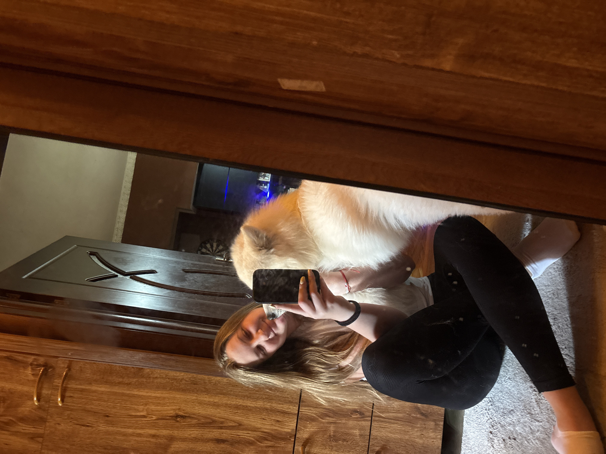 A woman is seen in the reflection of a wardrobe mirror. She is holding a smartphone, apparently taking a selfie with a large, white, fluffy dog. The woman is wearing black leggings with white spots, a white shirt, and a black wristband. She has long, blonde hair. The wardrobe is made of wood, and a door with a decorative design is also visible in the reflection. The floor has a carpeted surface. The city is Unknown, Unknown.