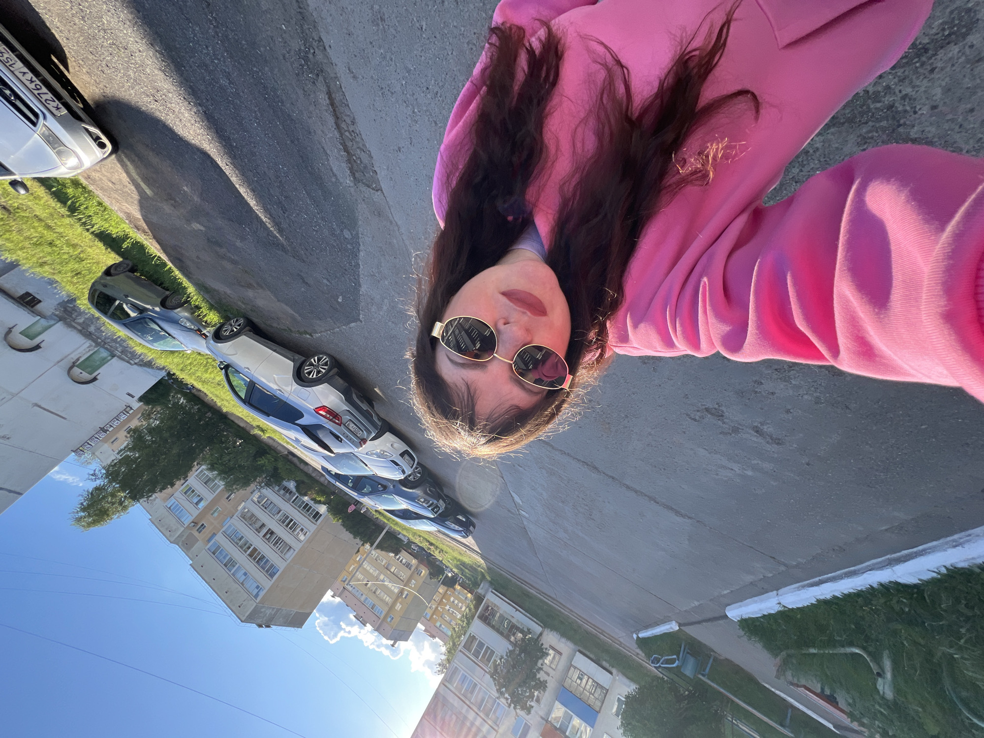 A woman wearing a pink hoodie and sunglasses is taking a selfie outdoors. In the background, there are several parked cars along a street adjacent to multi-story apartment buildings. The location is in Noviy, Russia, as indicated by the license plate on one of the cars with the Russian format. The sky is blue with some clouds, and the sun appears to be shining brightly. The buildings have a typical Soviet-era architectural style. There is greenery visible in the scene.